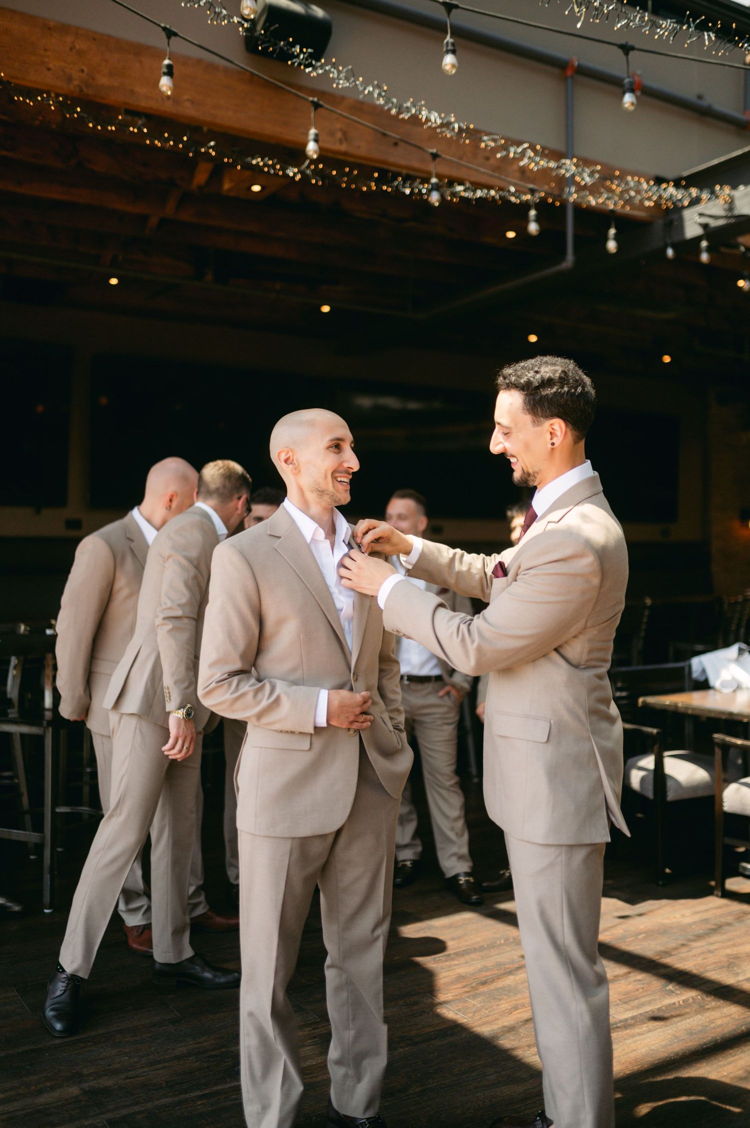 Wedding party in beige suits helping each other with final preparations indoors.