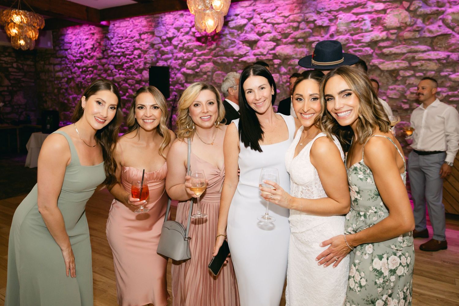 Wedding guests in pink and neutral dresses pose together against a stone wall with purple lighting.