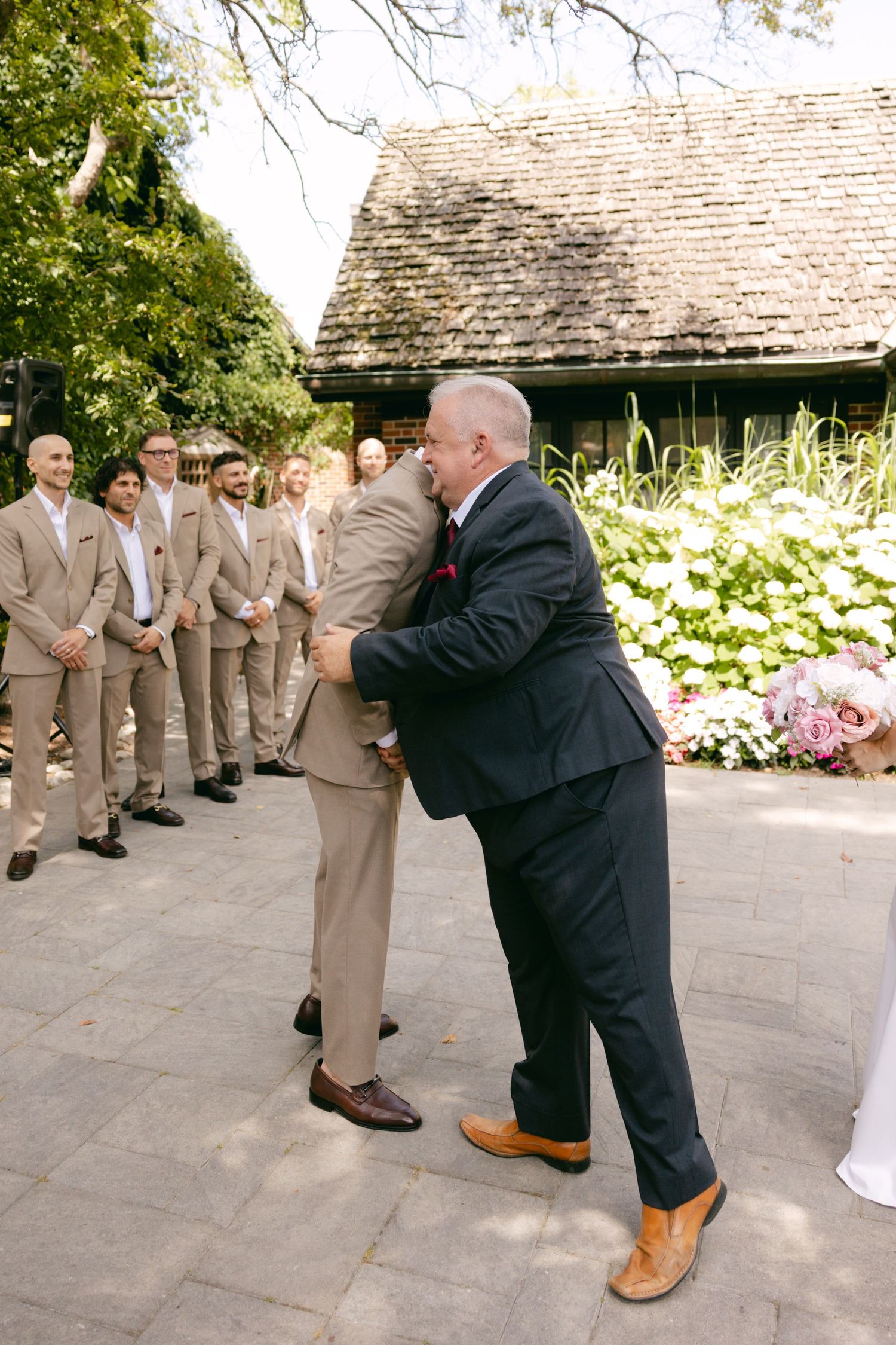 Wedding photographer captures candid moments of the bridal party in tan suits on a stone patio.