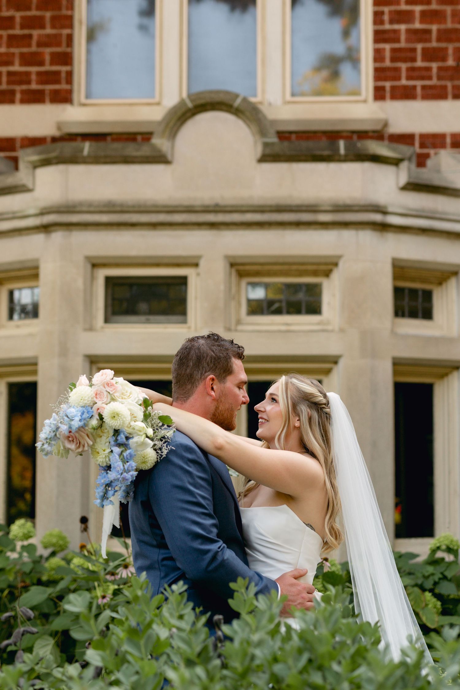 Wedding couple embraces in front of a historic brick building with landscaped gardens.