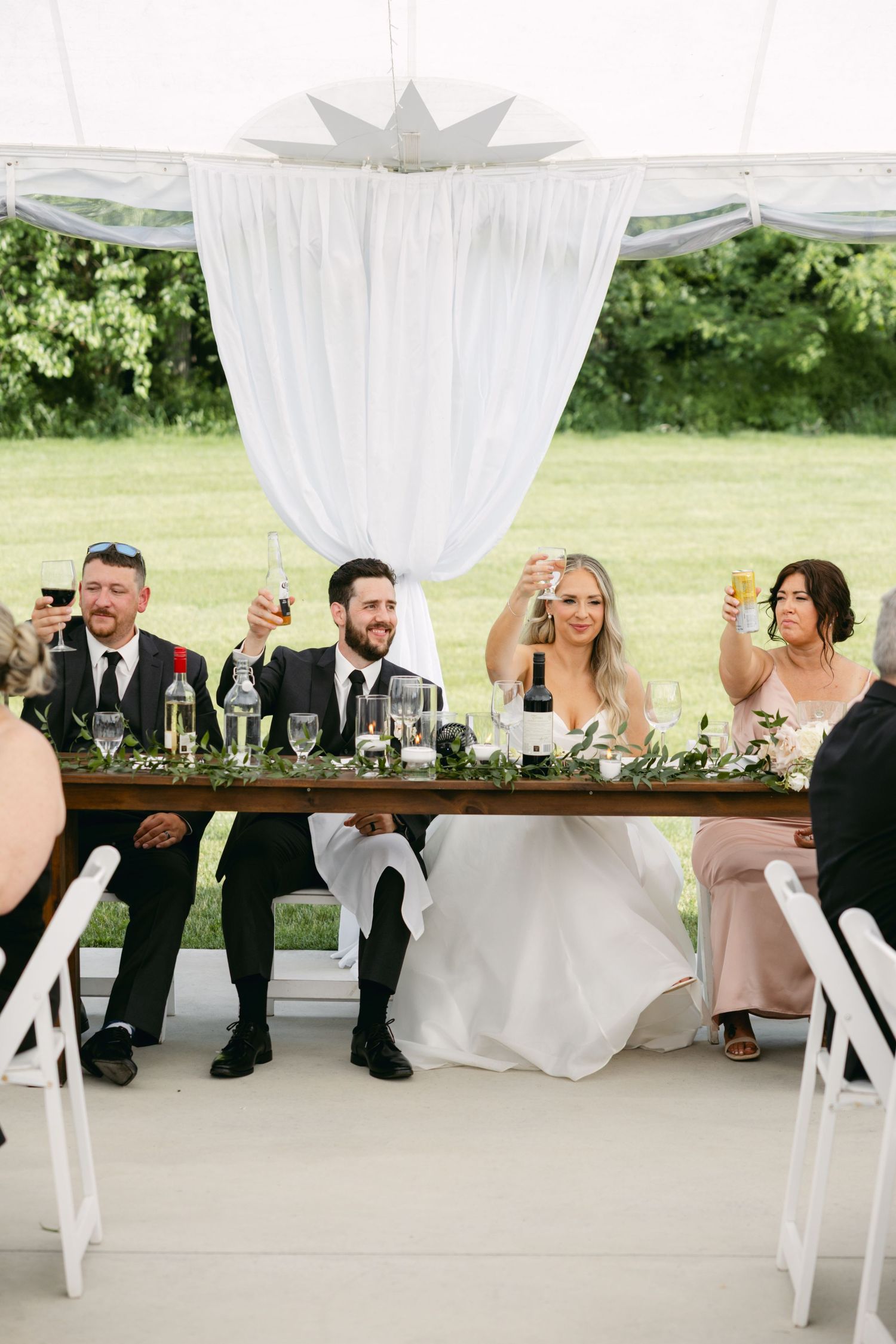 Wedding party members seated at a long table decorated with greenery under a white tent.