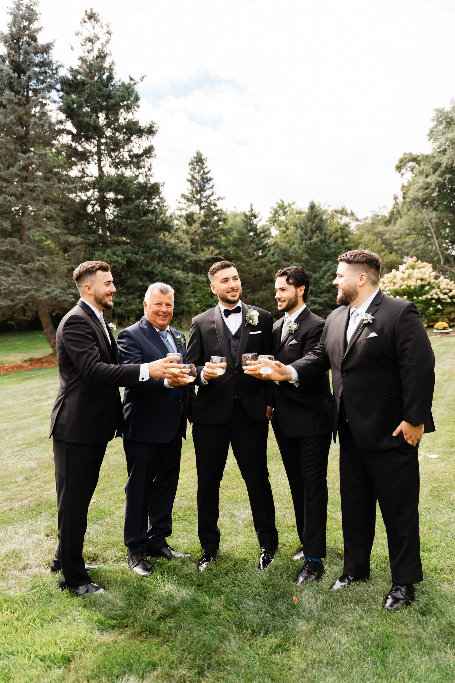 Groomsmen in formal attire toasting and celebrating together on a grassy hill.