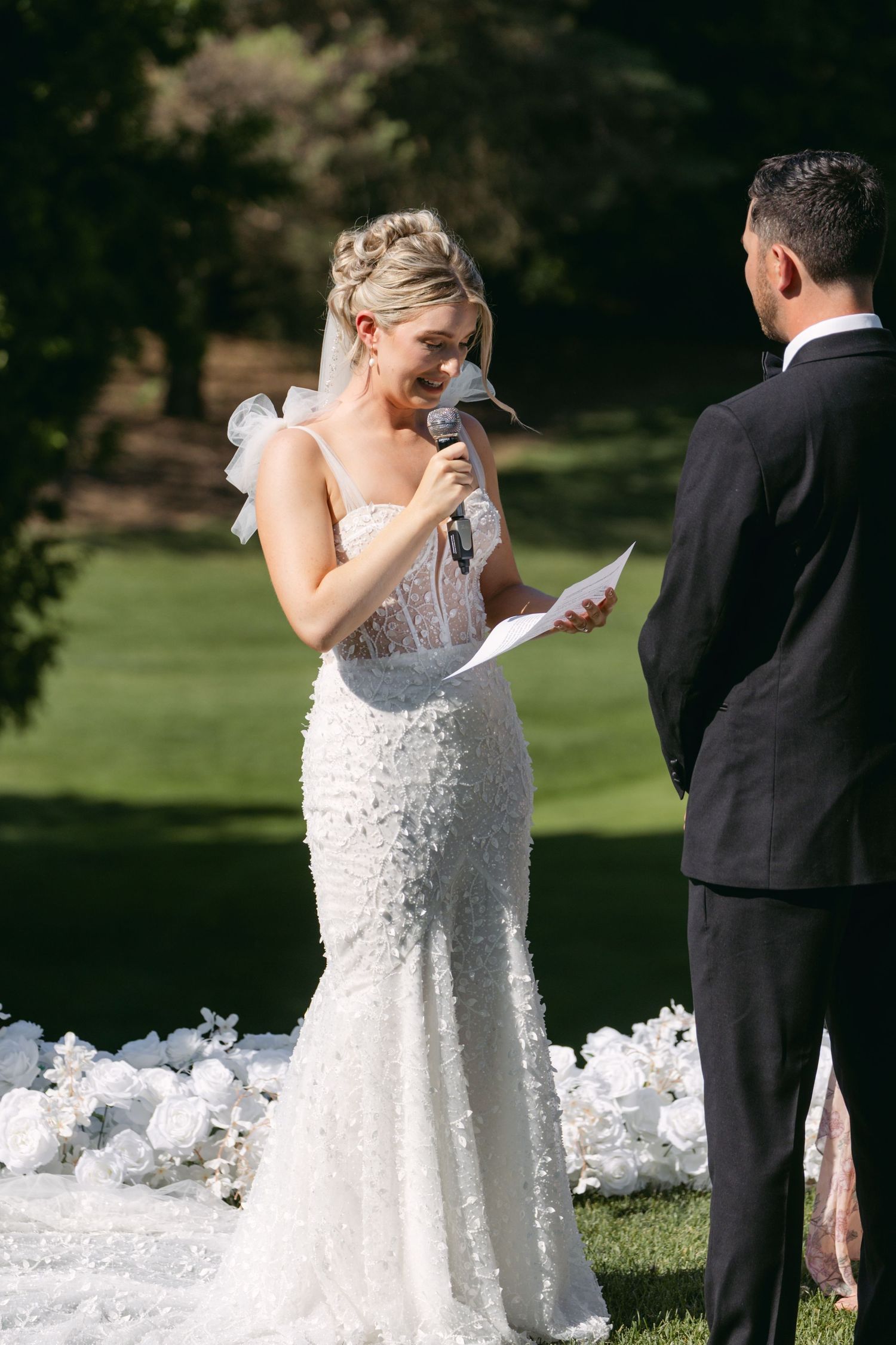 Bride in white lace dress reading vows during an outdoor ceremony.