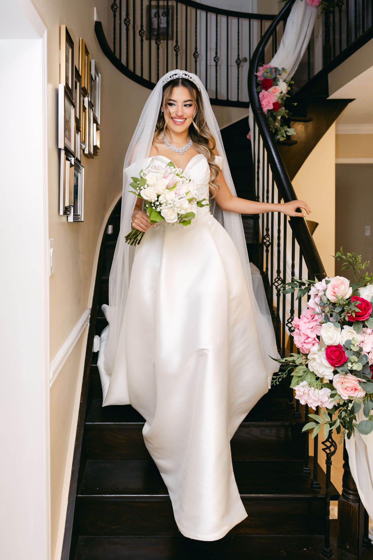 Bride in elegant white gown descending staircase decorated with pink and red flowers.