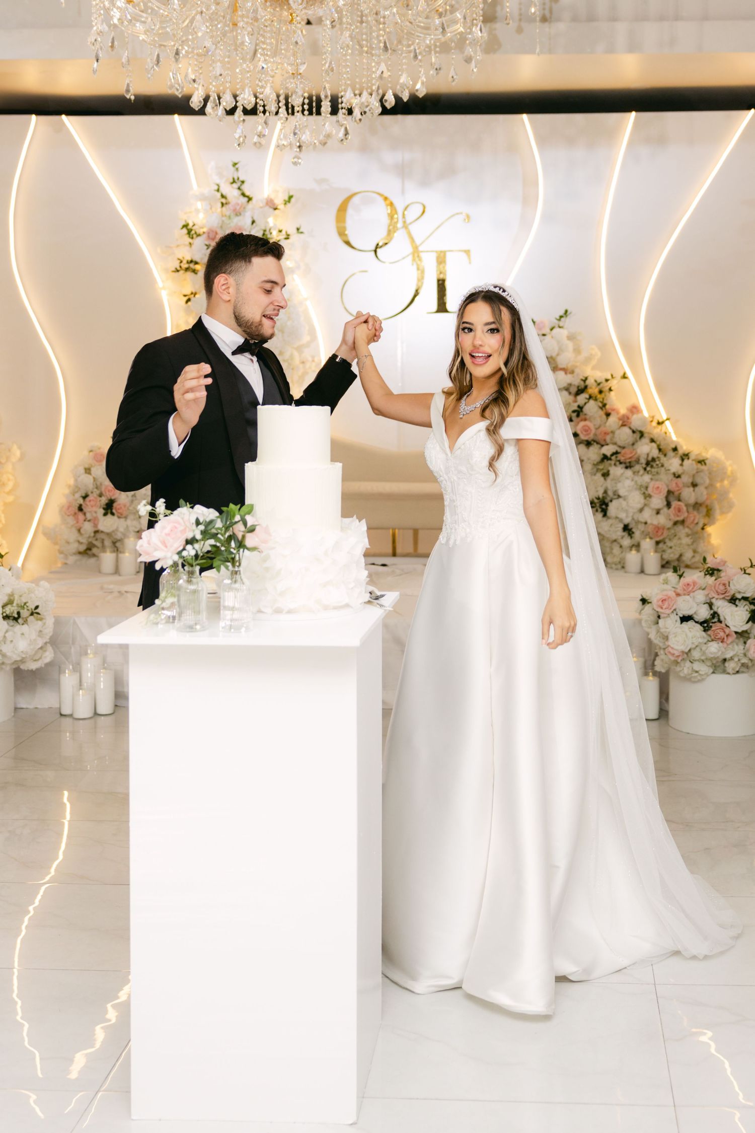 Newlyweds celebrate by their elegant white wedding cake under a crystal chandelier.