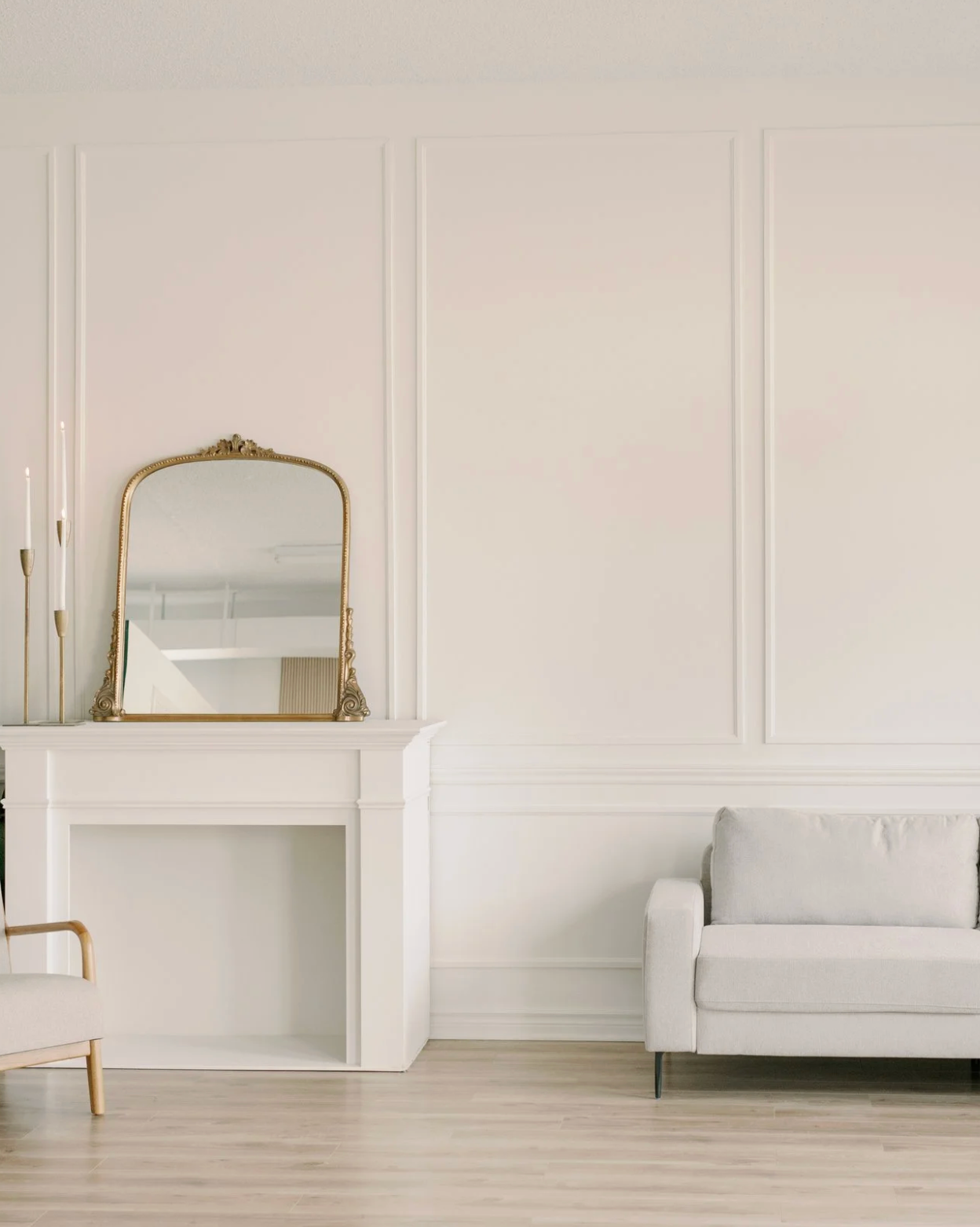 Elegant white living room with ornate gold mirror above fireplace, light gray sofa and classic wall paneling details.