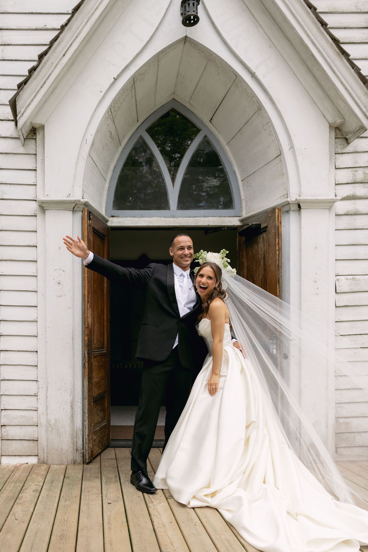 Joyful bride and groom posing at the entrance of a white wooden chapel after their wedding ceremony.