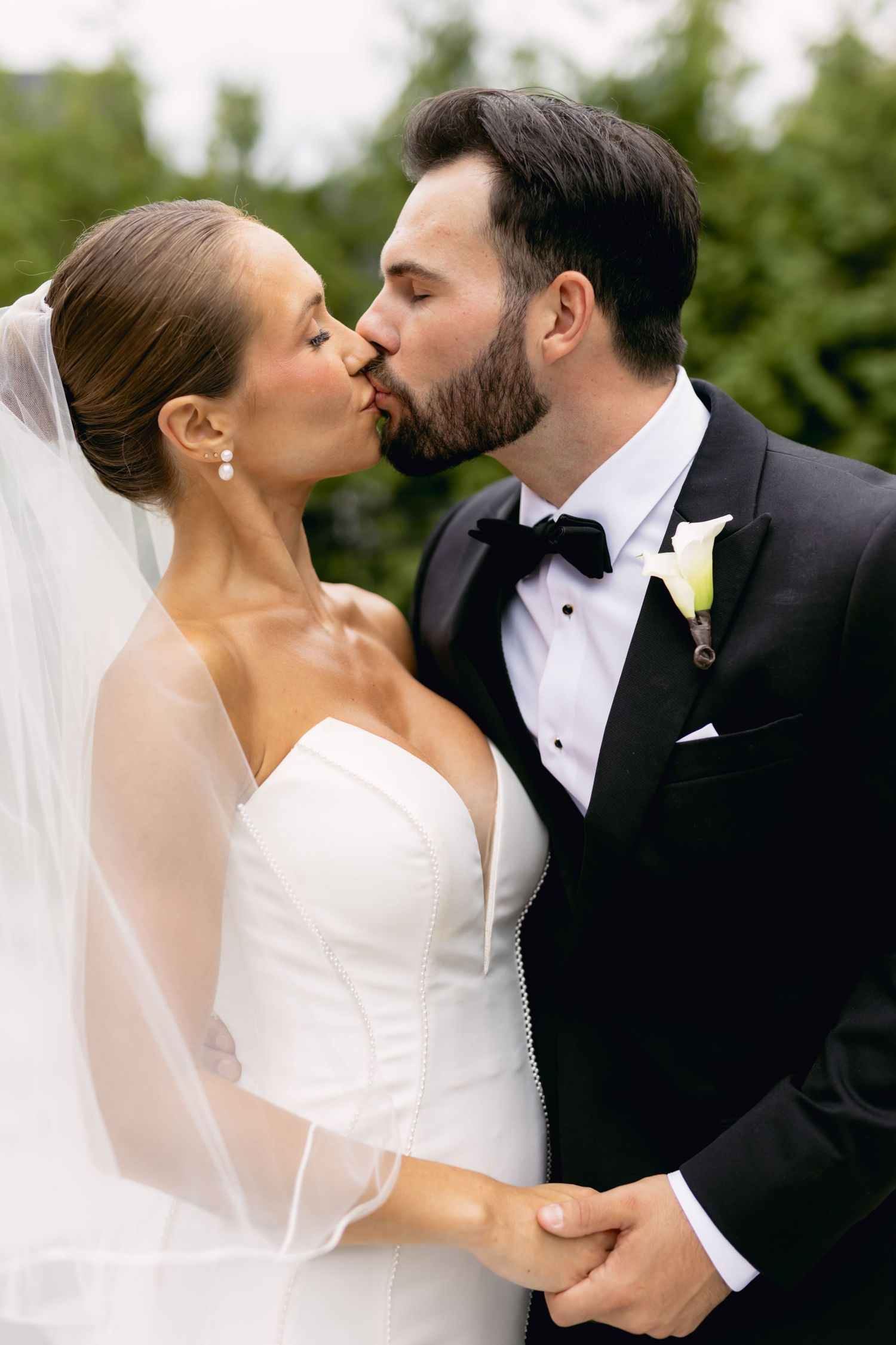 Bride and groom share a kiss outdoors, groom in black tuxedo with bow tie, bride in strapless white gown with veil.