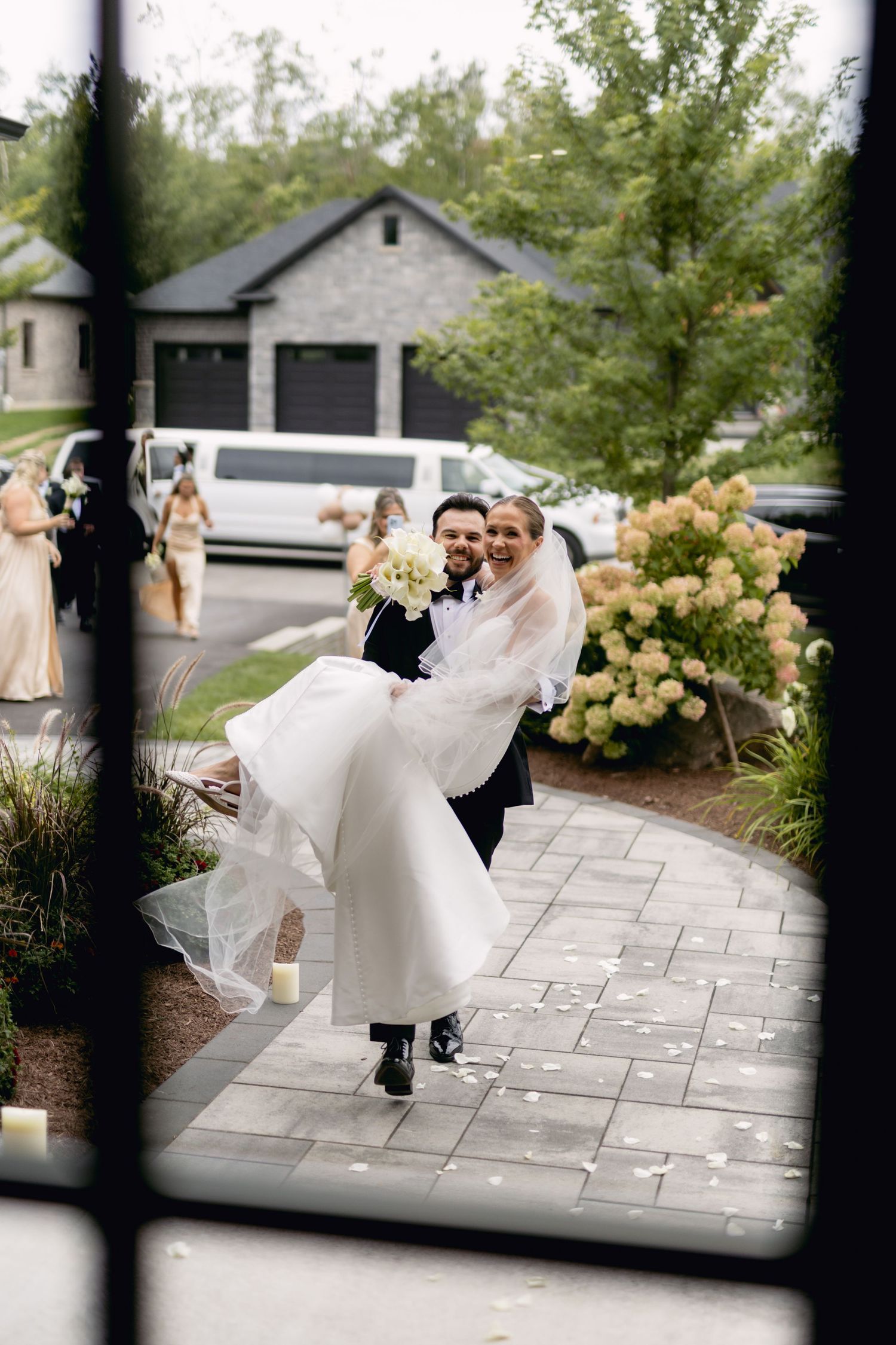Groom carries bride in white gown and veil along paved path with wedding party and limo in background.