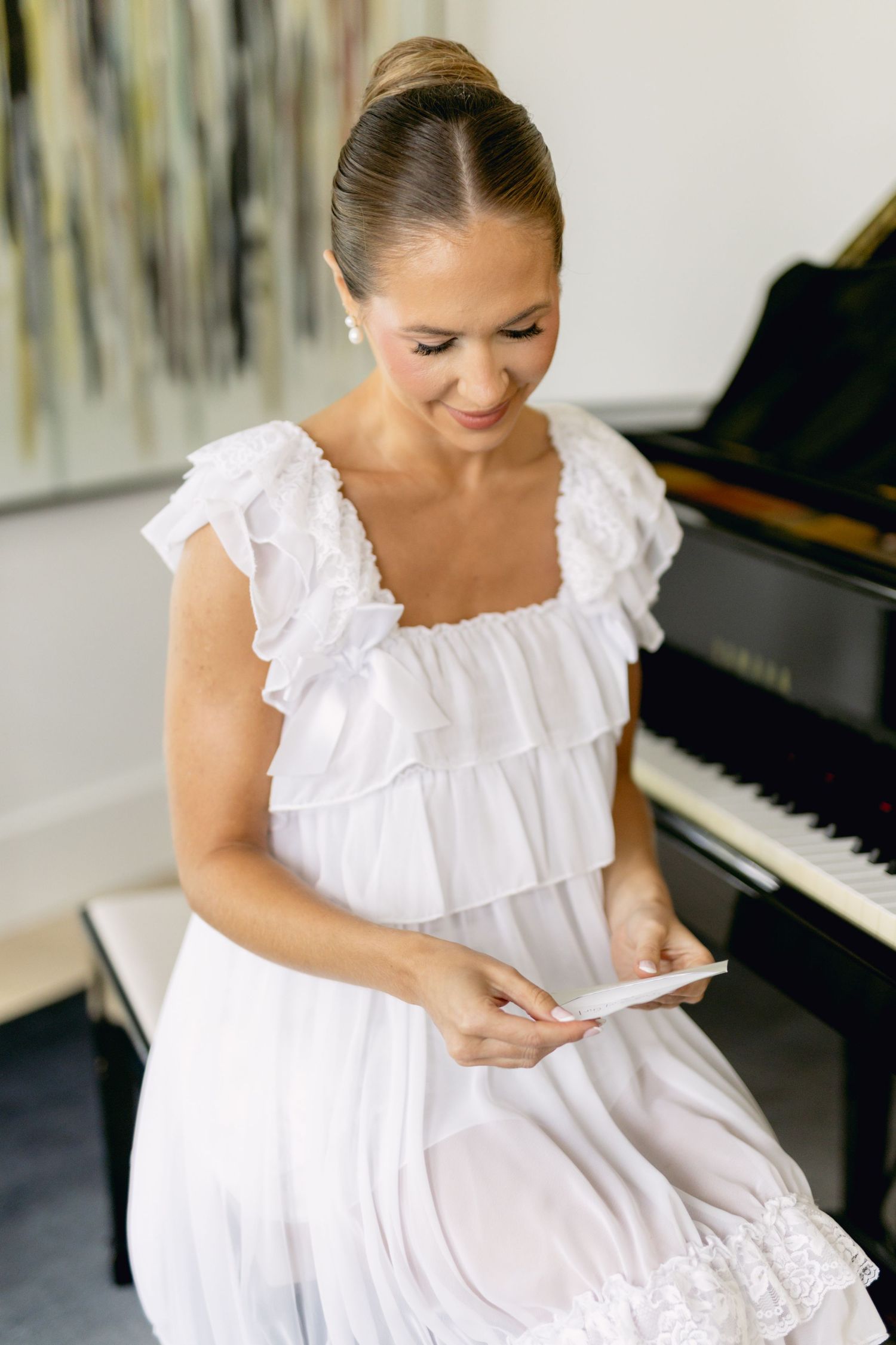 Bride in white ruffled dress reading a note while seated near a grand piano.