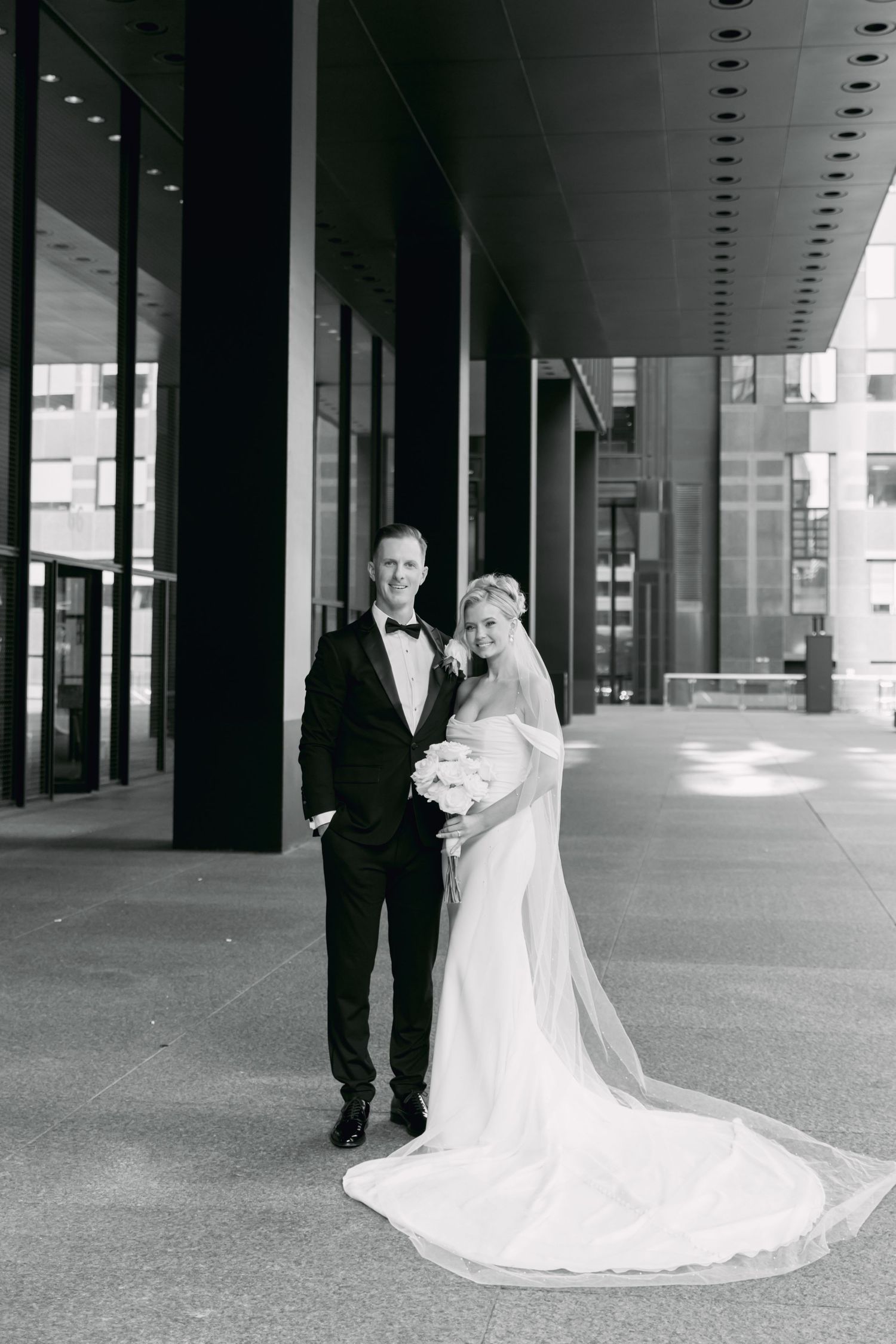 Black and white portrait of elegant bride and groom posing outside modern city building with train flowing.