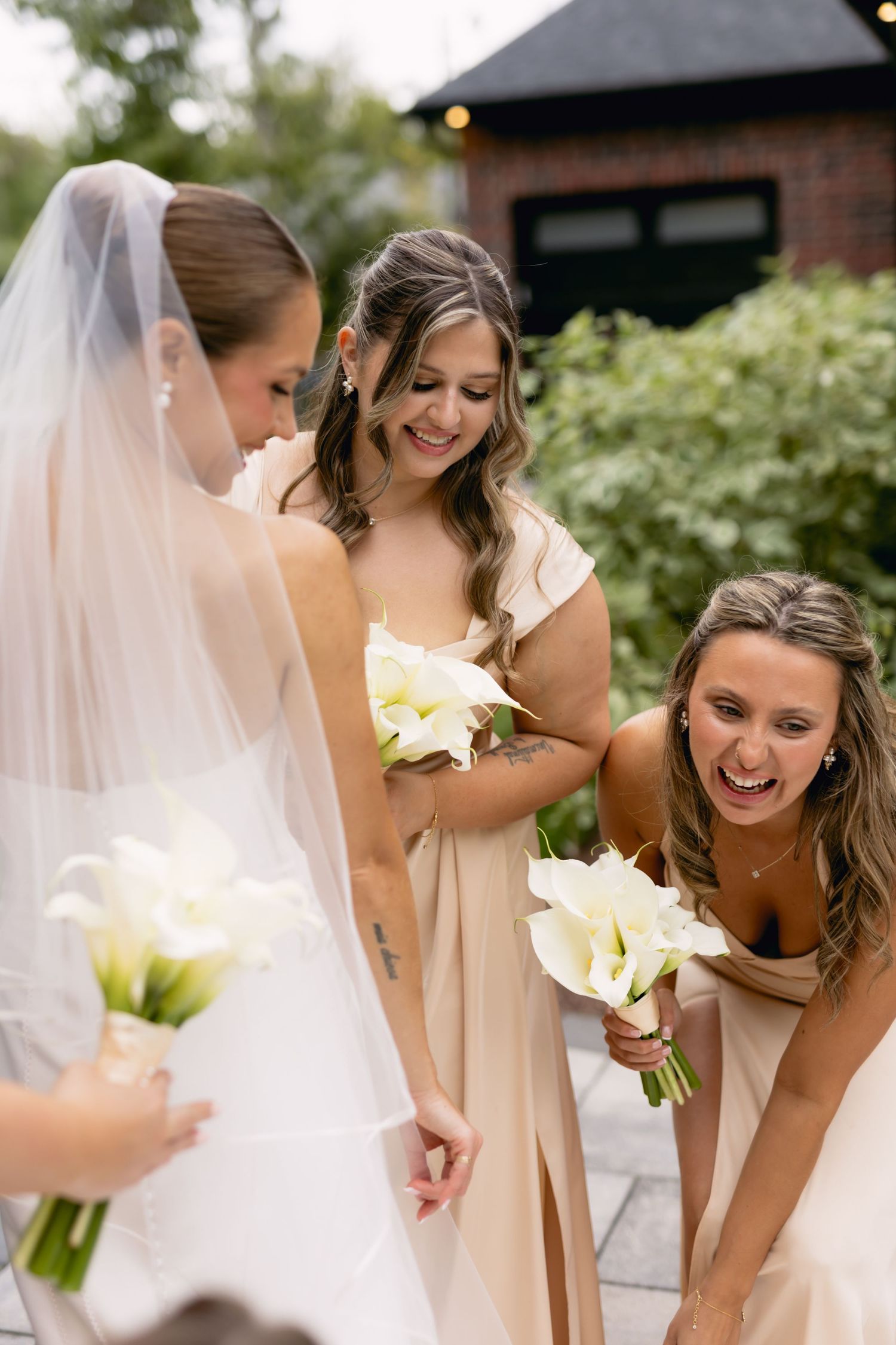 Bride in white gown and veil stands with two laughing bridesmaids in champagne dresses holding white calla lily bouquets.