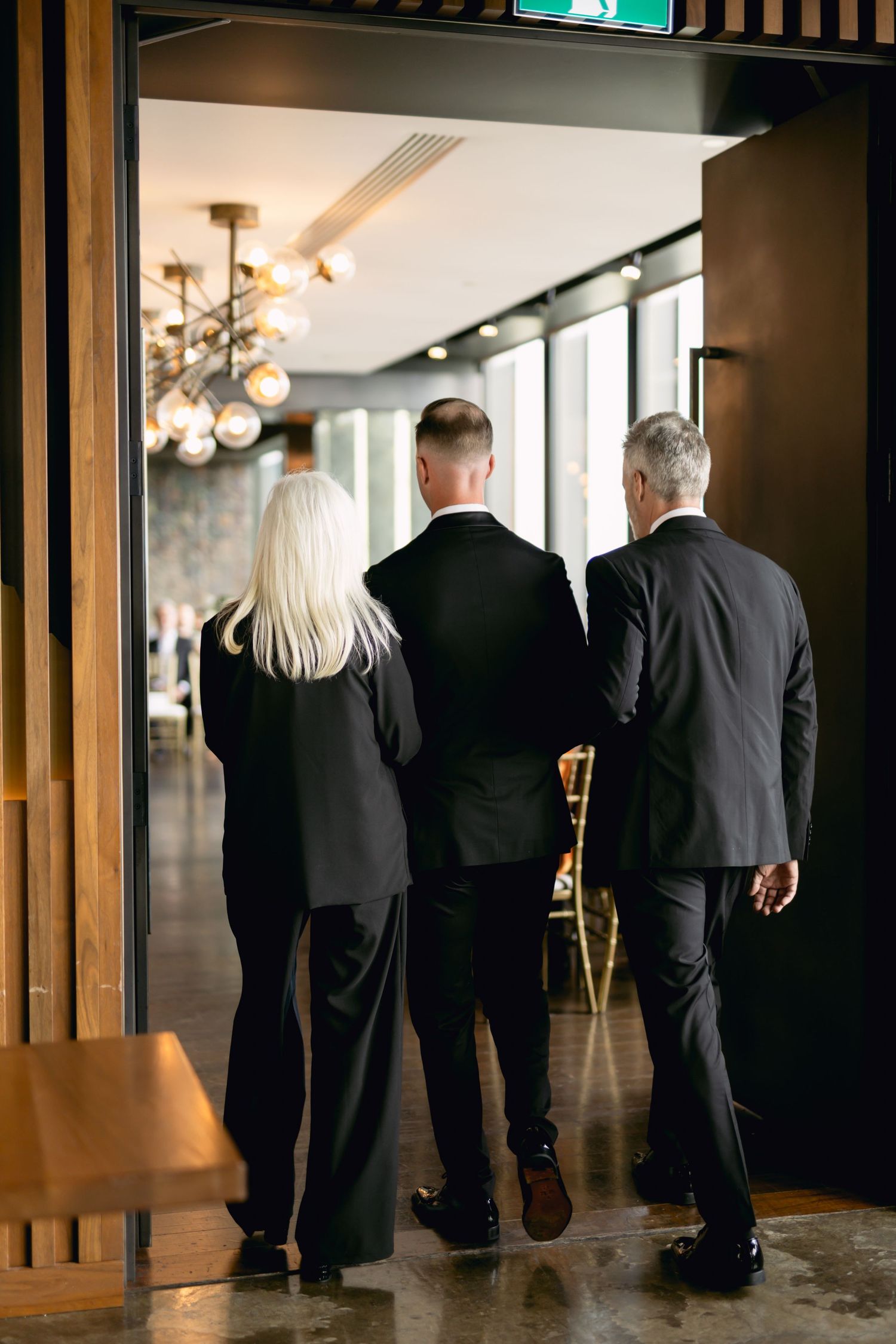 Three elegantly dressed wedding guests in black attire walking into a modern reception venue with chandelier lighting.