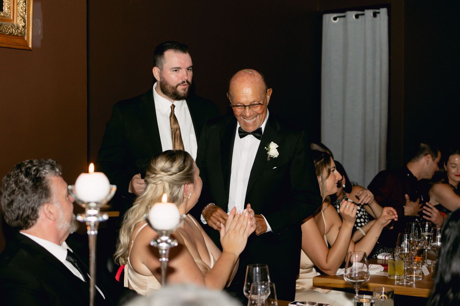 Older gentleman in tuxedo greets seated wedding guests during elegant candlelit reception dinner.