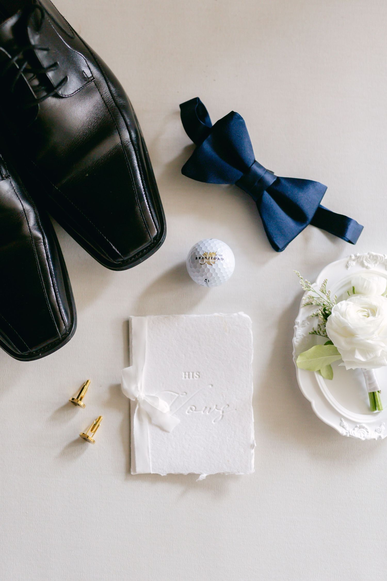 Wedding accessories including dress shoes, bow tie, golf ball and white floral boutonniere arranged on white surface.