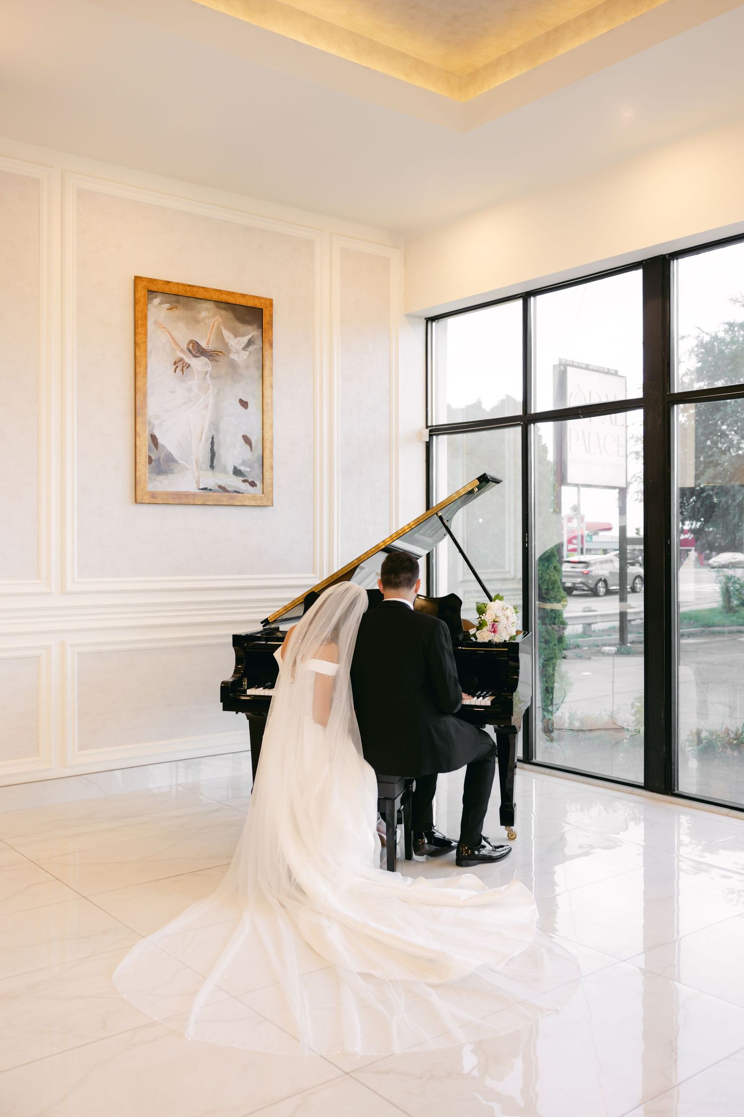 Bride in flowing white gown poses near grand piano in modern bright interior with natural light.