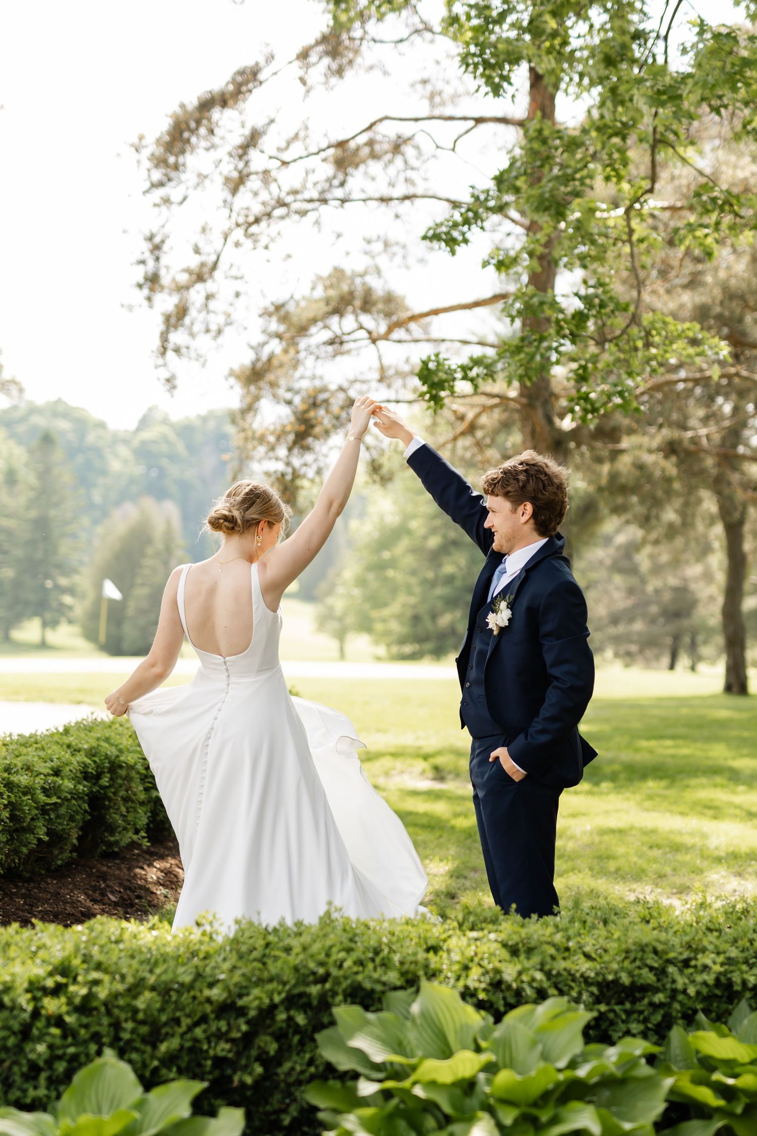 Playful wedding couple dancing in garden with manicured hedges and trees.