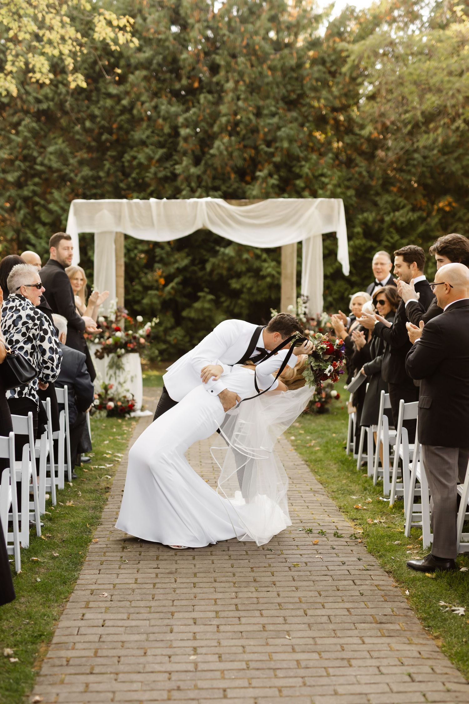 Wedding celebration on stone pathway with white chairs and draped arch during autumn ceremony.