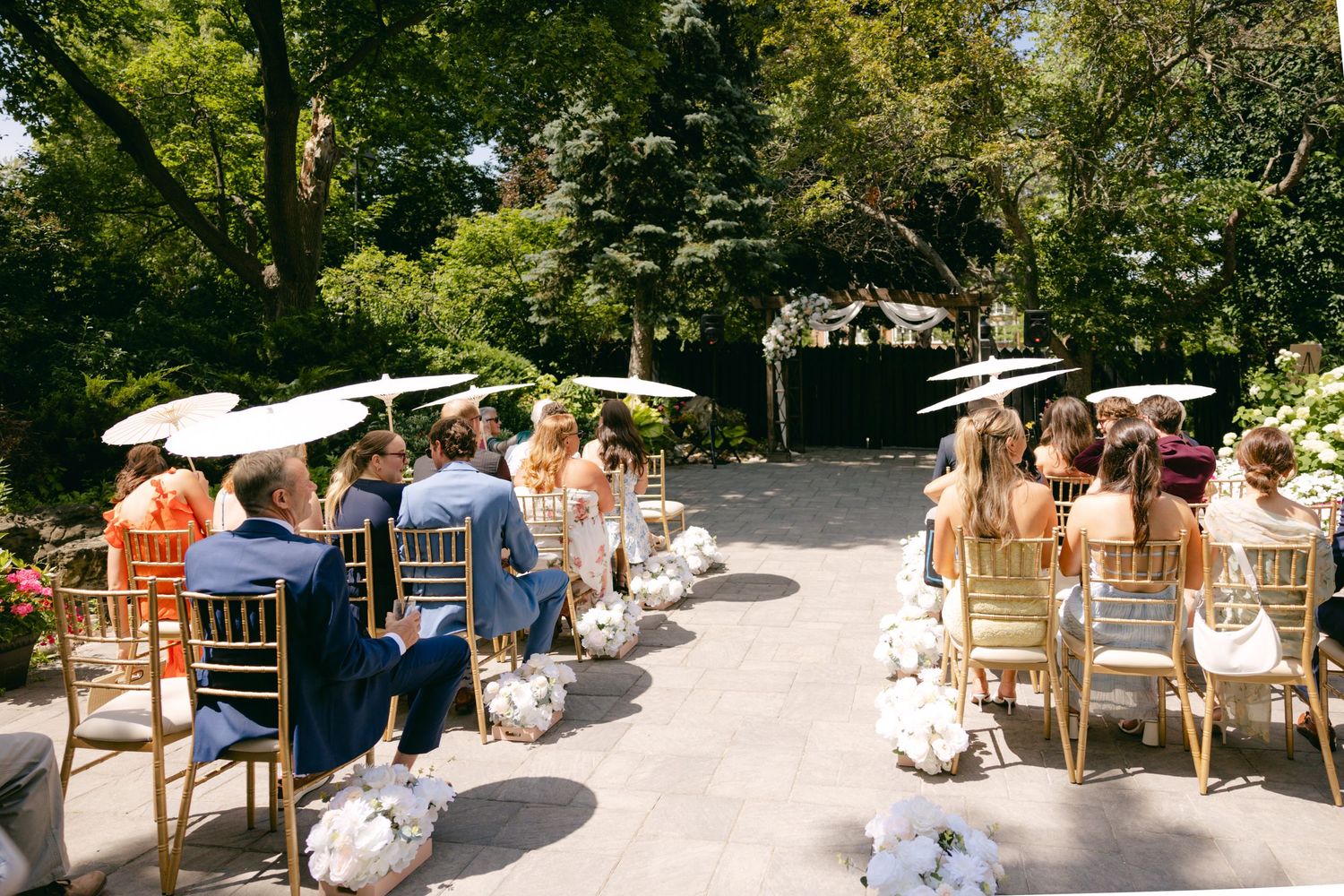 Wedding guests seated with white parasols during an outdoor garden ceremony.