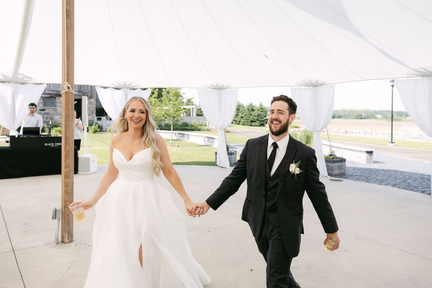 A joyful wedding couple enters their reception tent holding hands with bright smiles.