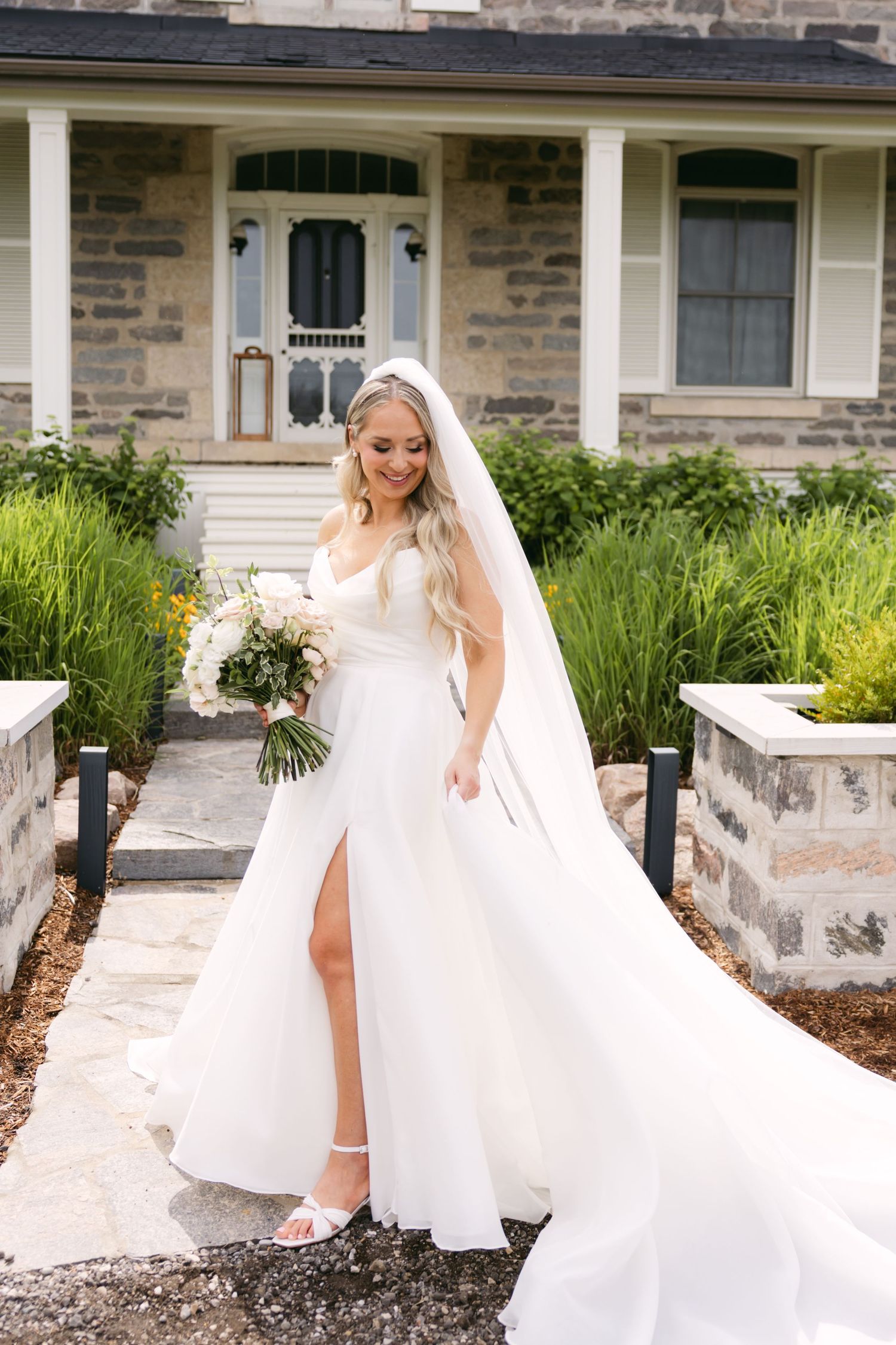 Bride in flowing white gown shows off her dress near stone building with white columns.
