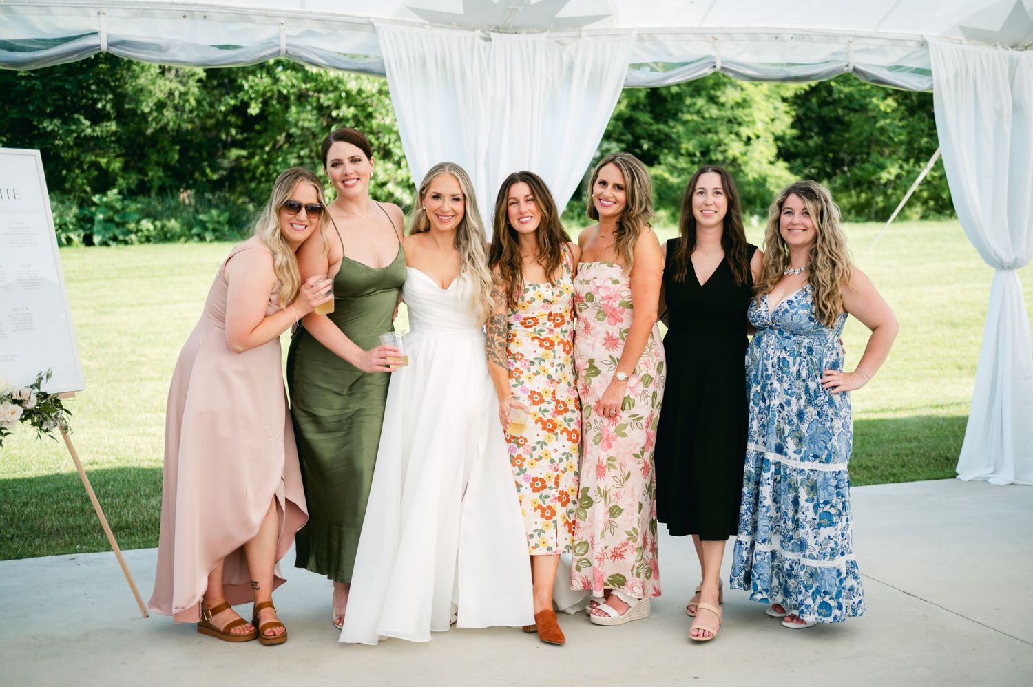 Wedding party members in colorful dresses pose together under a white tent at an outdoor celebration.