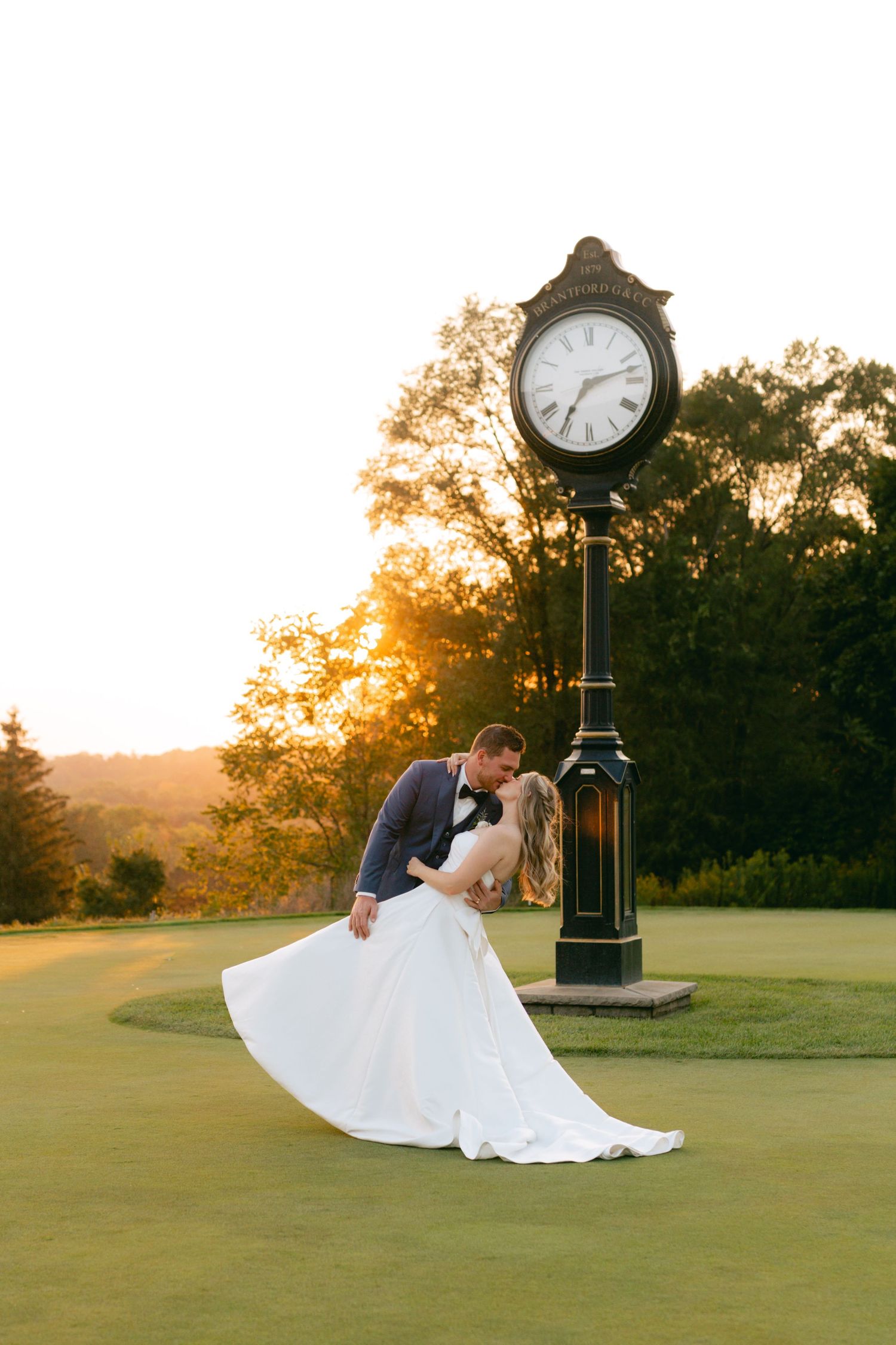 Newlyweds share a romantic moment by a vintage clock on a golf course at sunset.