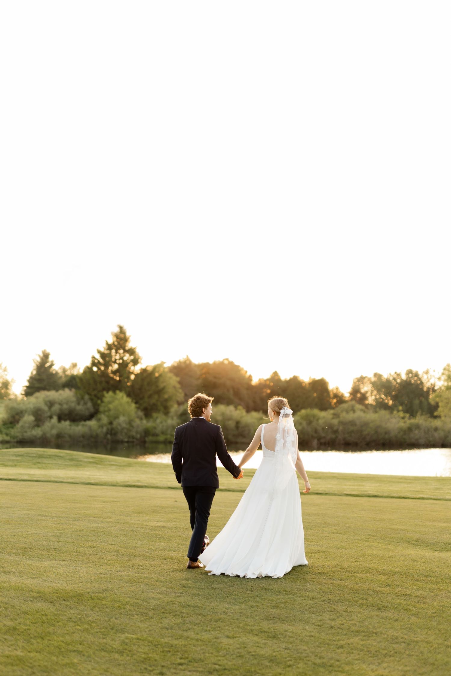 Bride and groom stroll hand in hand across an open field at sunset.