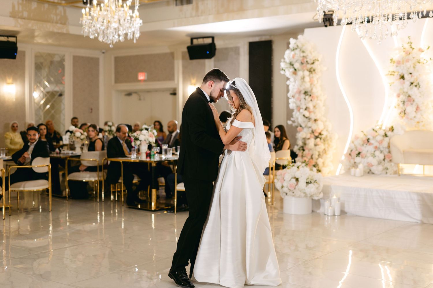 Couple dancing romantically under ornate chandeliers in a luxurious white reception venue.