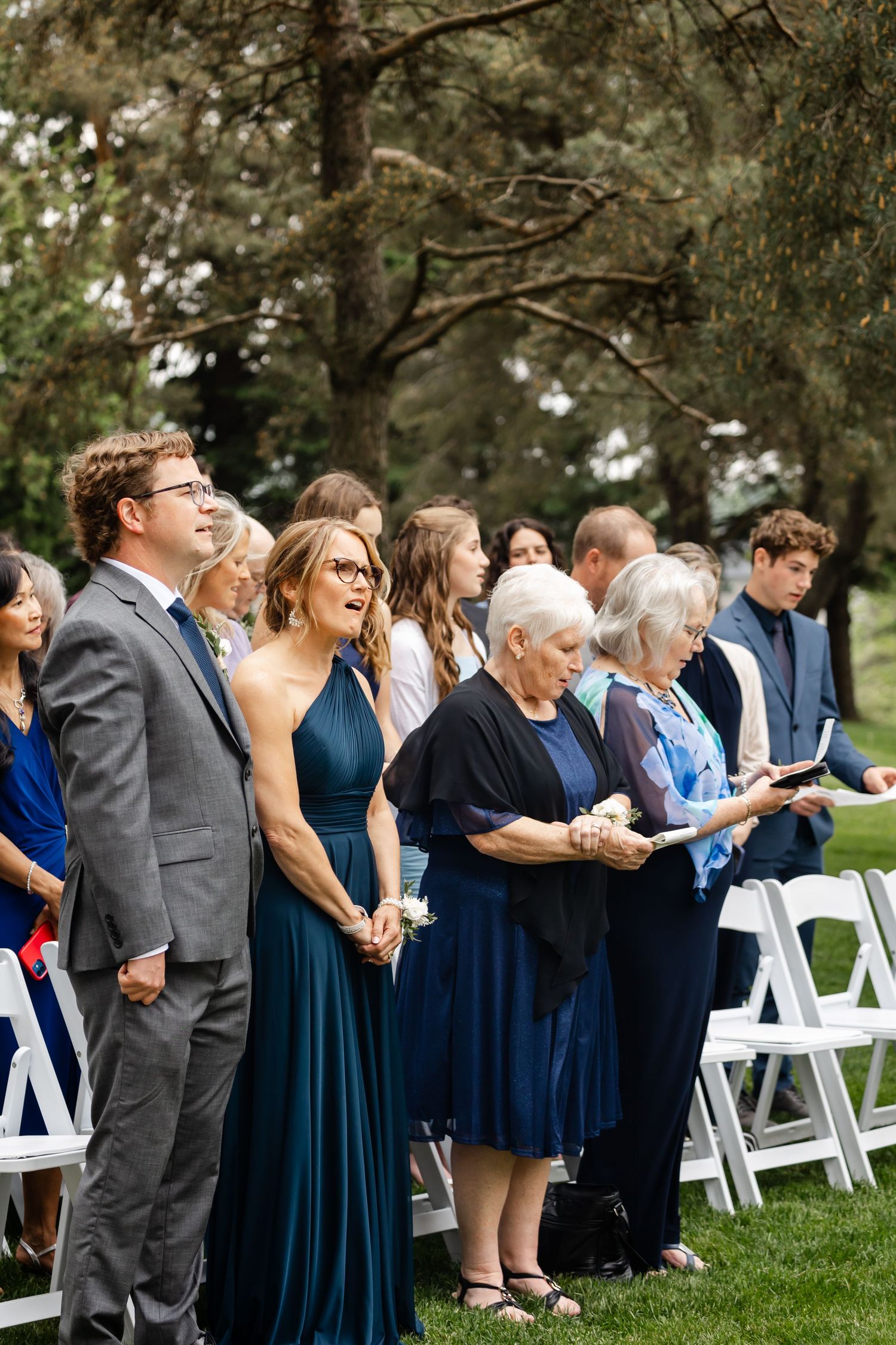 Wedding guests singing from hymnals during outdoor ceremony with evergreen trees in background.