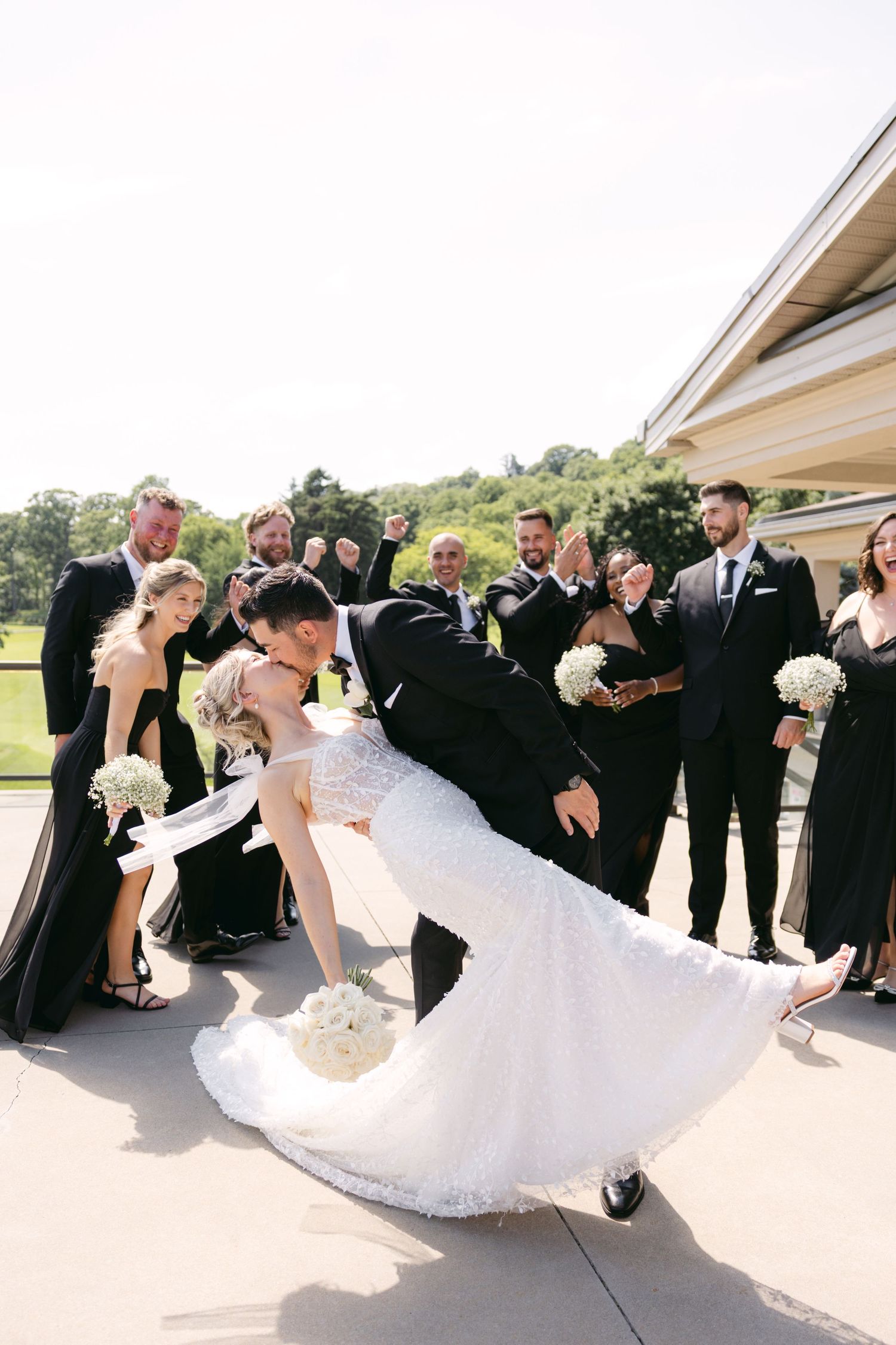 Wedding party performs fun dip pose with bride and groom in center on sunny outdoor patio.