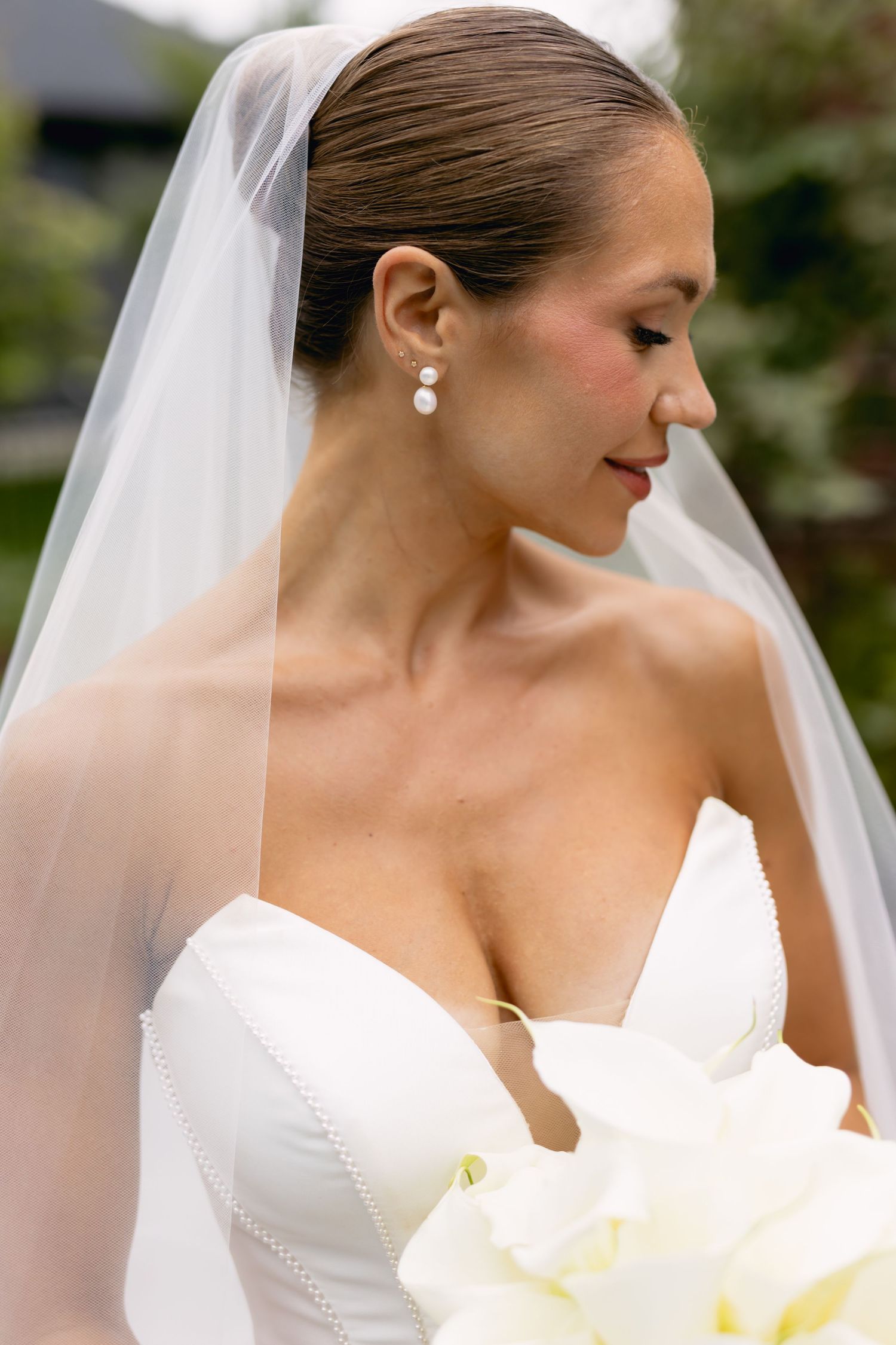 Close-up of bride in strapless white gown and veil wearing pearl drop earrings holding bouquet.