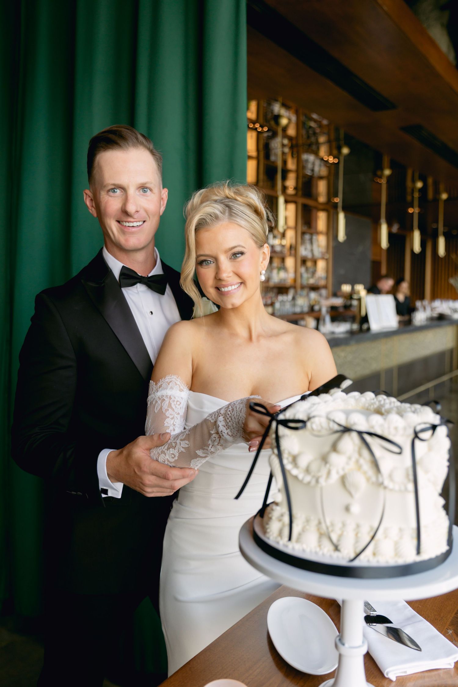 Bride and groom smile together while cutting elegant white wedding cake with black bow decoration.