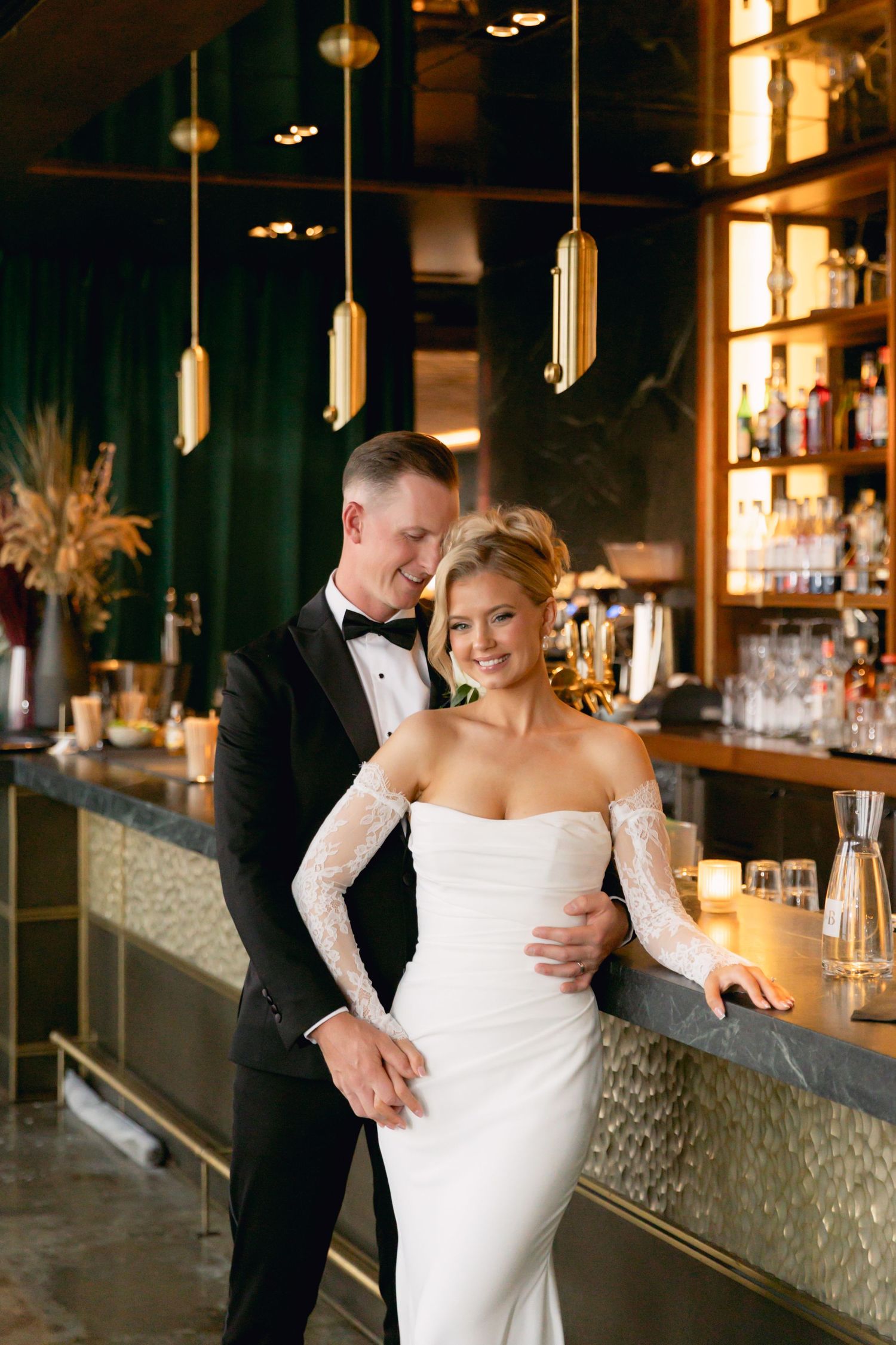 Smiling bride and groom embrace at elegant bar with warm gold lighting and green velvet backdrop.