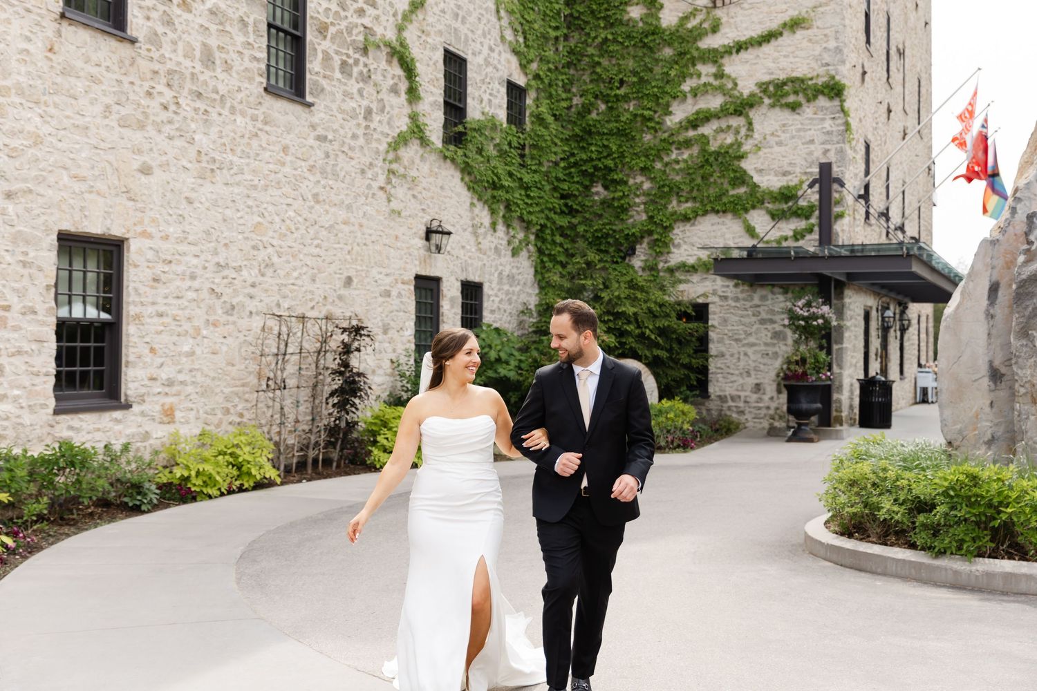 Bride and groom walk arm-in-arm past ivy-covered stone building at elegant wedding venue.