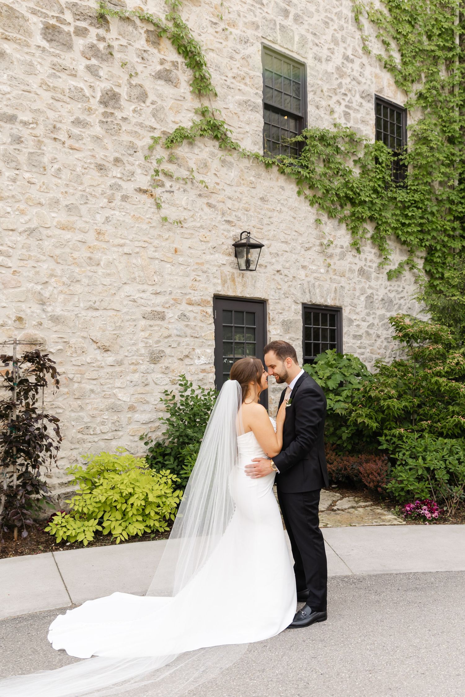 Bride and groom embrace tenderly in front of an ivy-covered stone building on their wedding day.