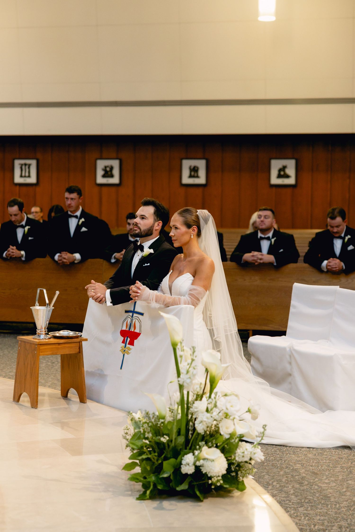 Bride and groom standing at church altar during wedding ceremony with groomsmen seated in background.