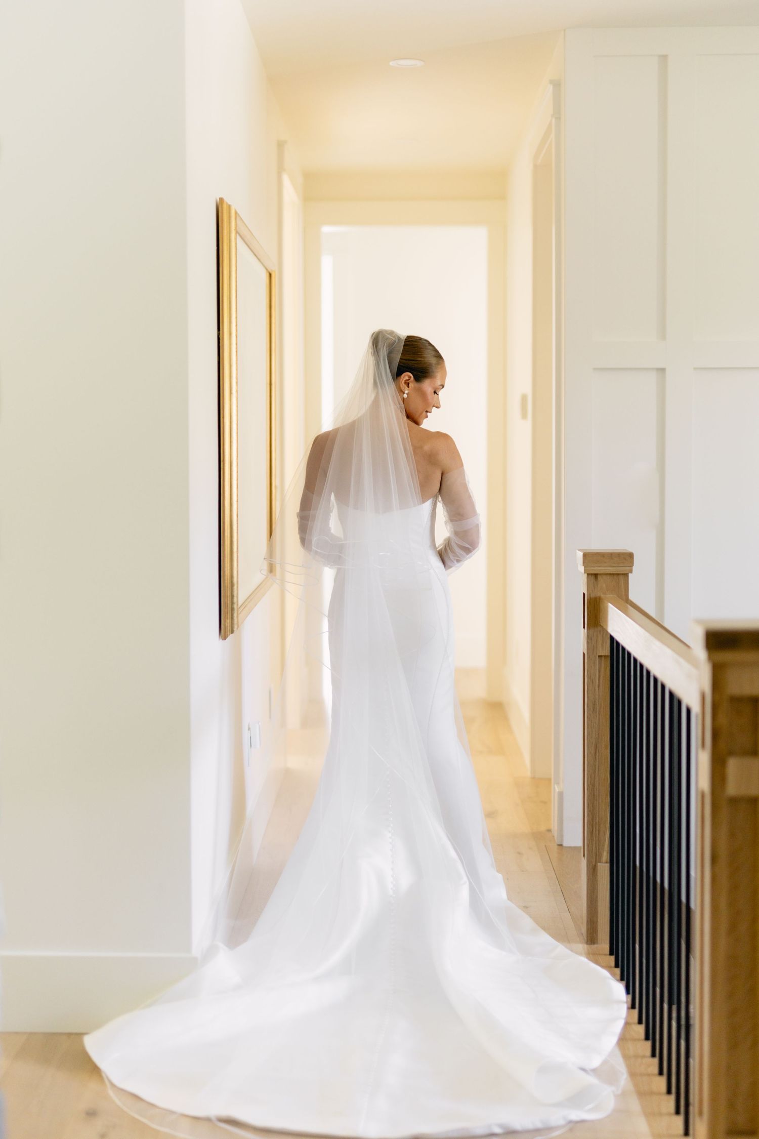 Elegant bride in fitted white gown with long veil standing in hallway viewed from behind.