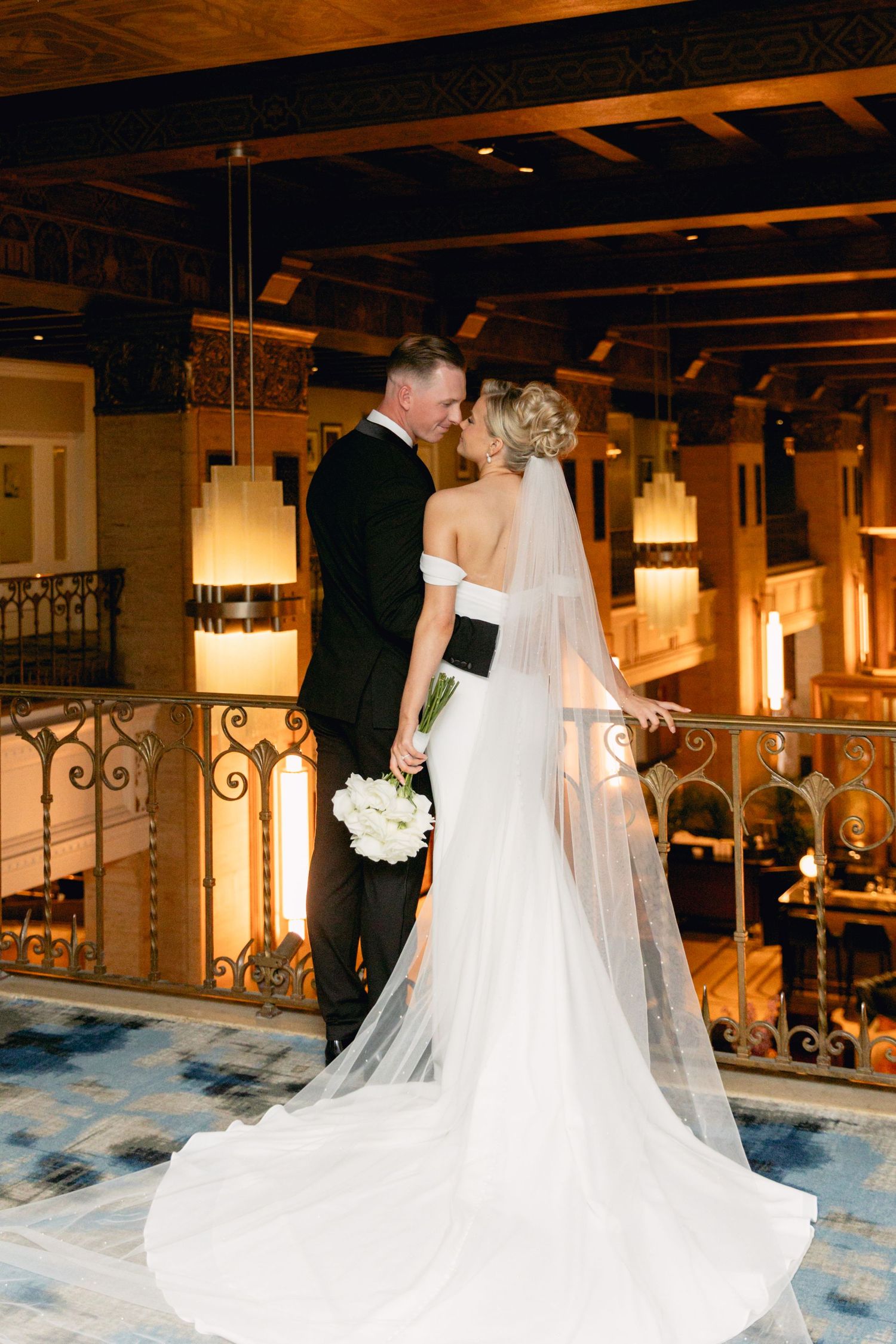 Bride and groom share a romantic moment on a balcony, her white off-shoulder gown and cathedral veil trailing behind.