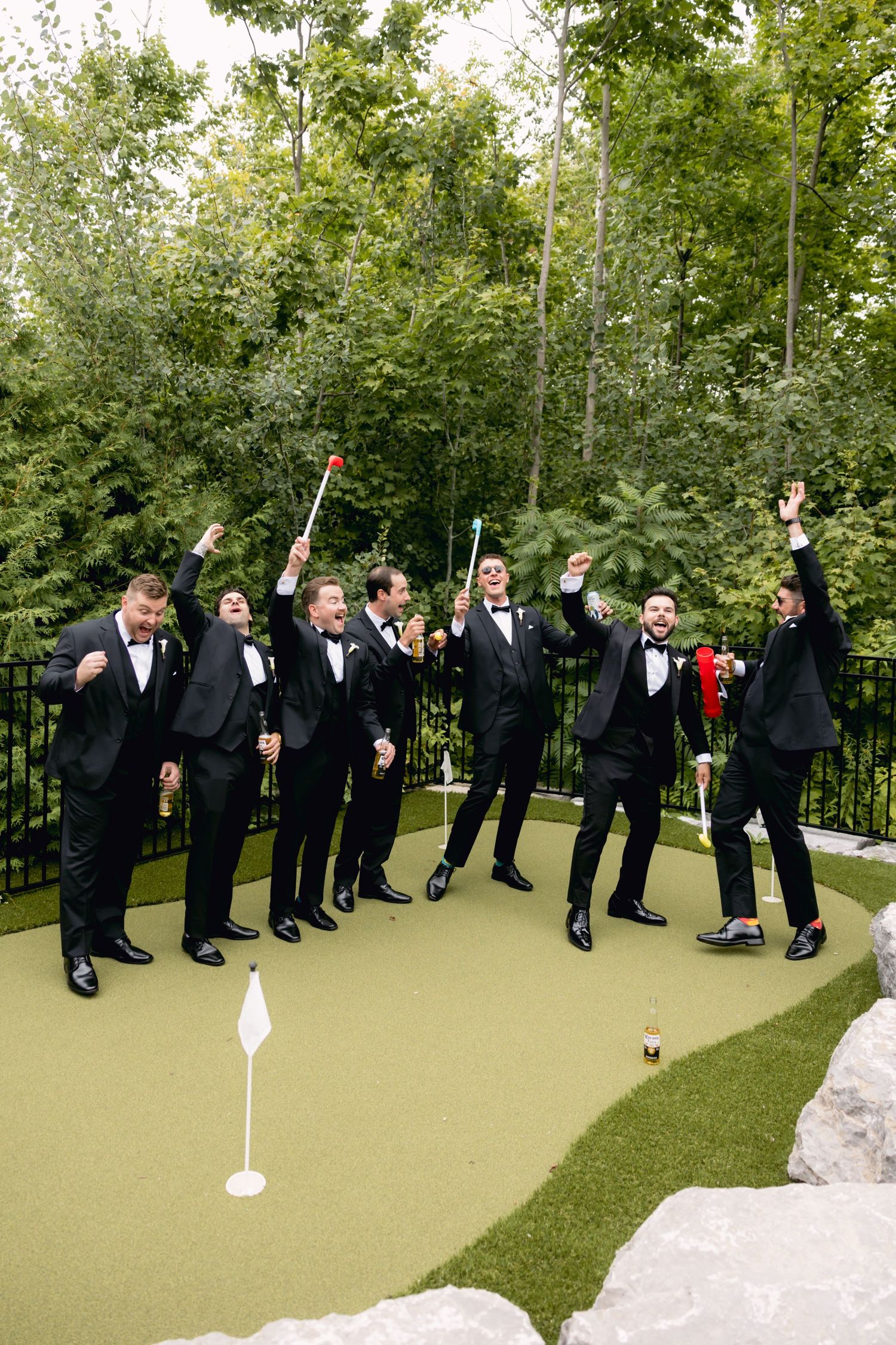 Groomsmen in black tuxedos celebrate on mini golf course, raising arms and golf clubs in excitement.