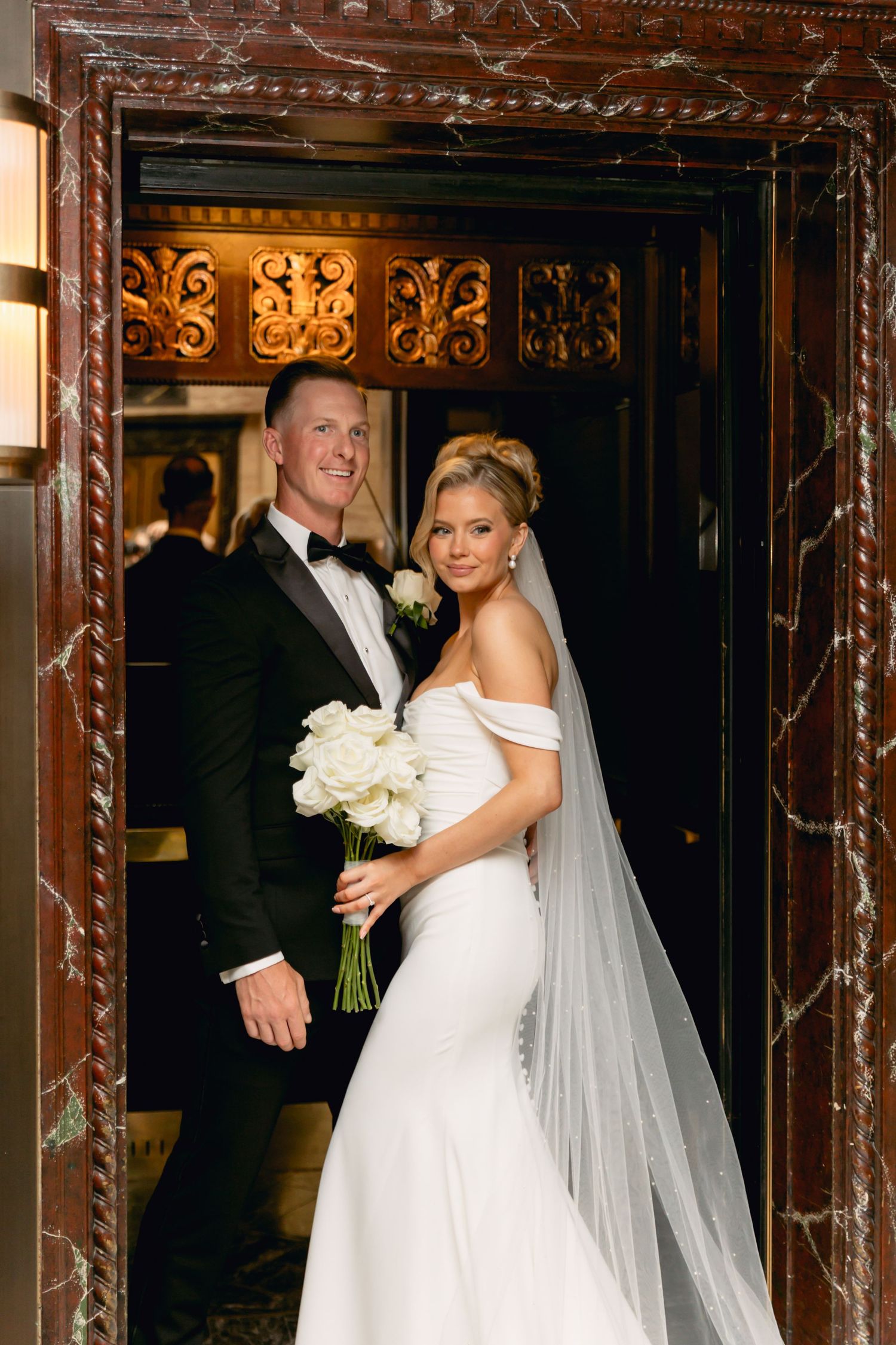 Smiling bride and groom pose in ornate doorway, bride holding white rose bouquet in fitted gown with veil.