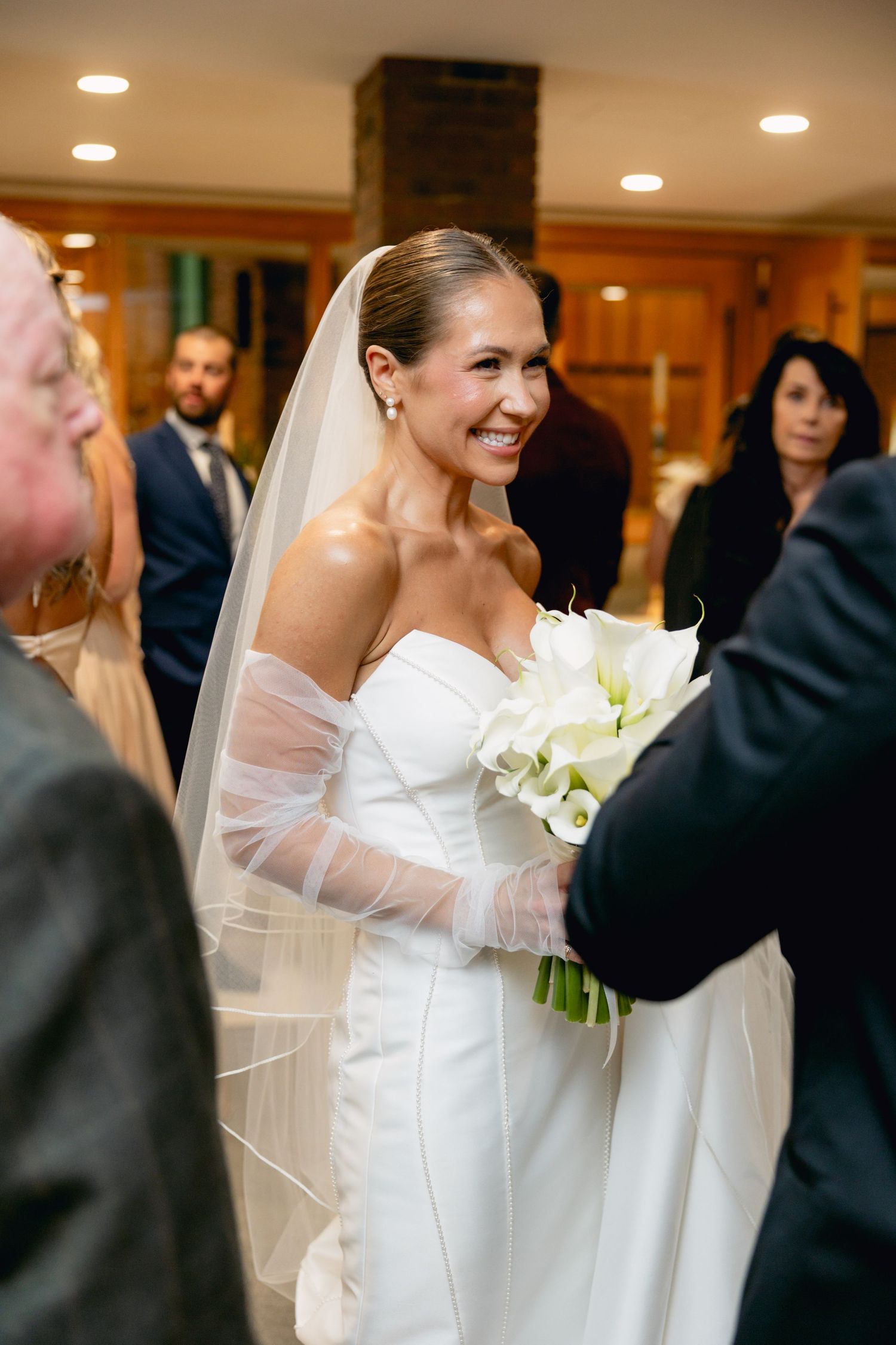 Radiant bride in strapless white gown and veil smiling while holding calla lily bouquet indoors.