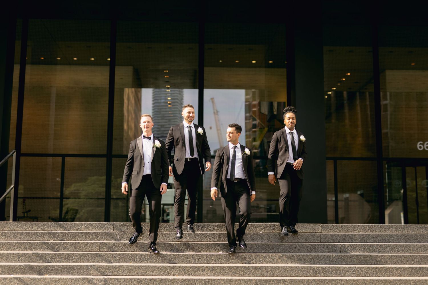 Four groomsmen in suits descend outdoor steps of modern glass building at night.