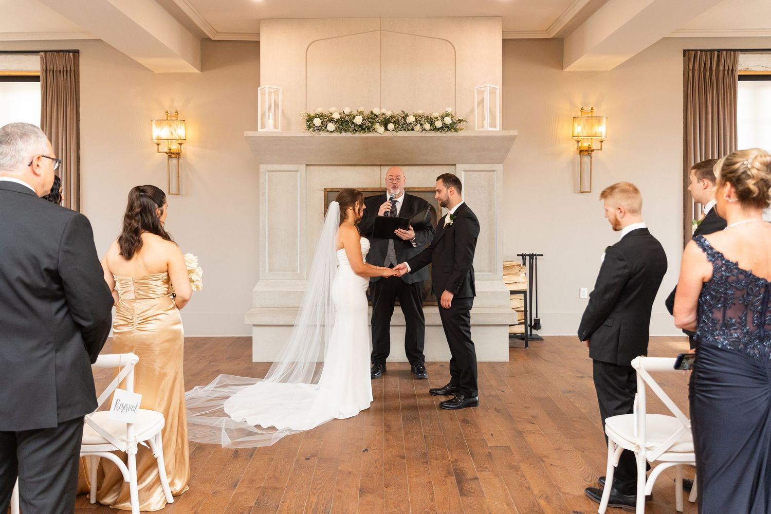Bride and groom exchange vows at an elegant indoor ceremony before a white fireplace with florals.