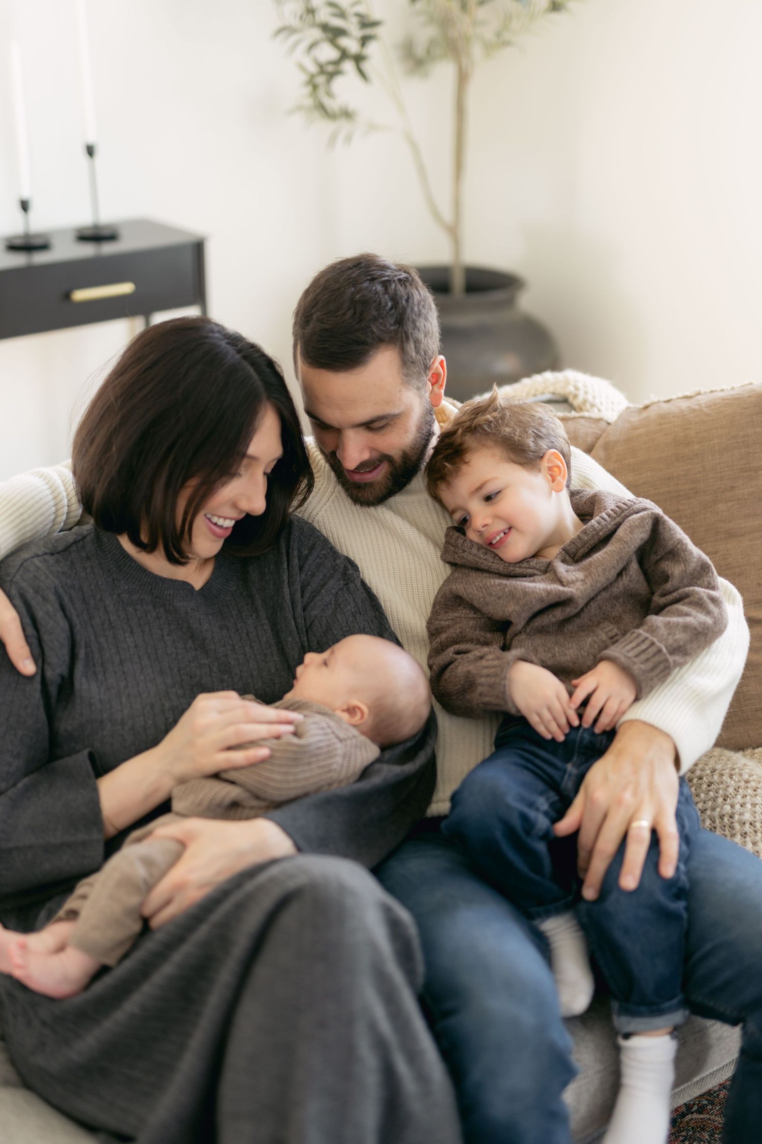 Happy family of four sitting on sofa, parents smiling at newborn baby while toddler boy looks on warmly.