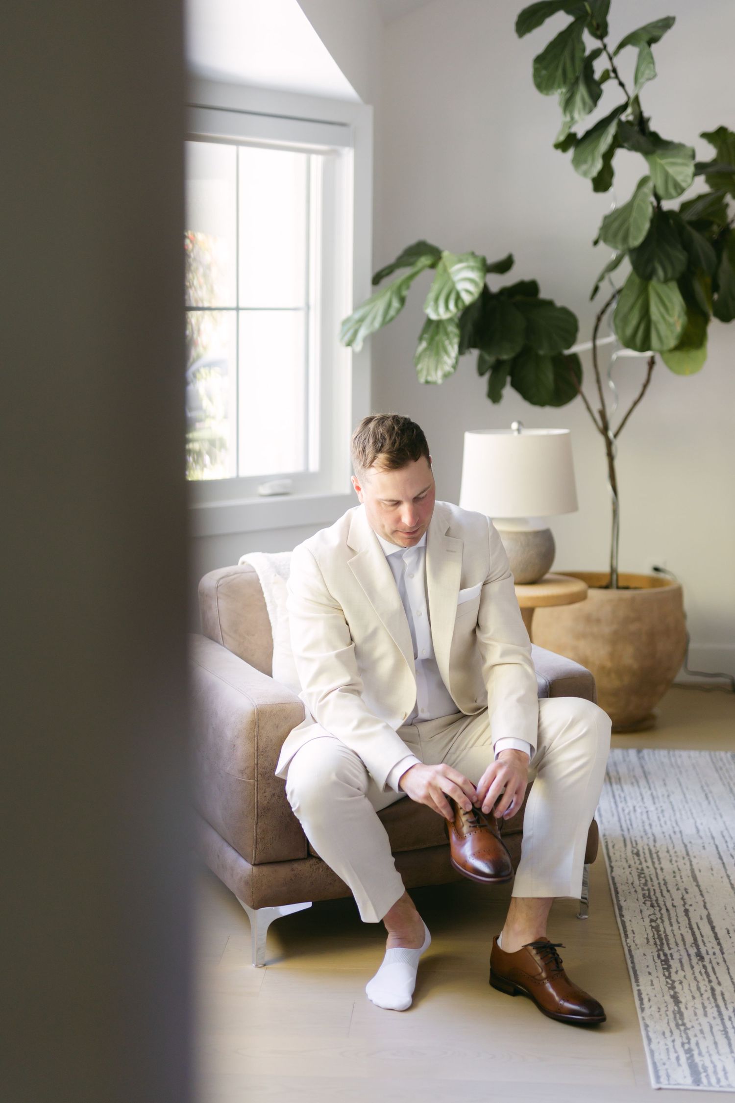 A person in a cream colored suit sits in a bright modern room with a fiddle leaf fig plant.