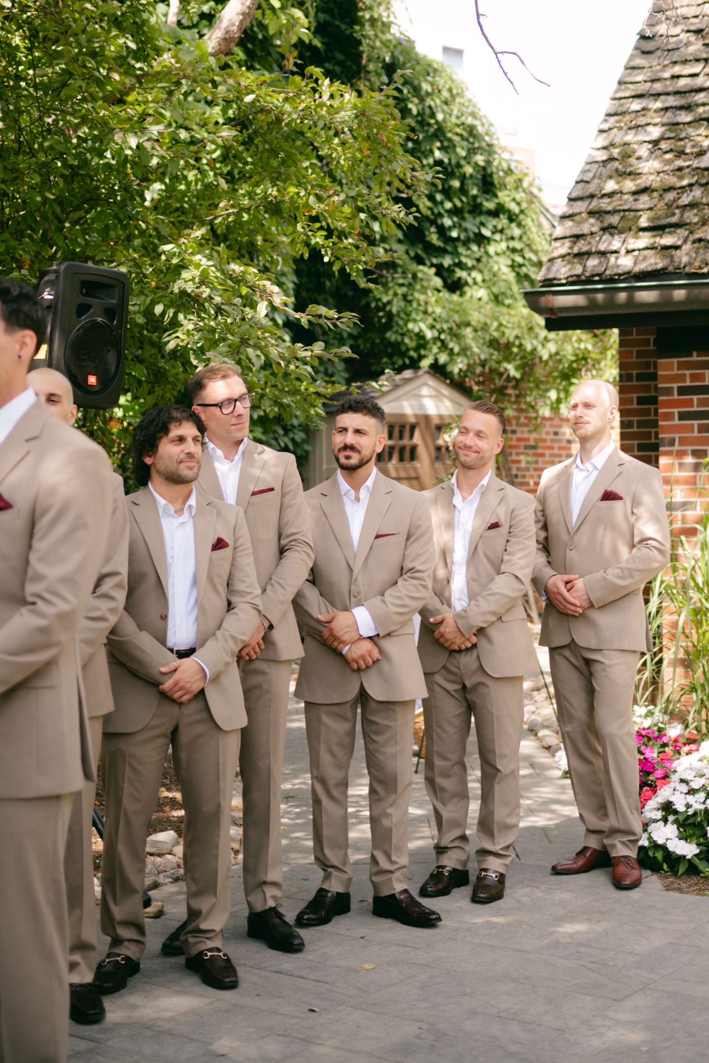 Groomsmen in tan suits pose together in a line against a brick wall and garden backdrop.