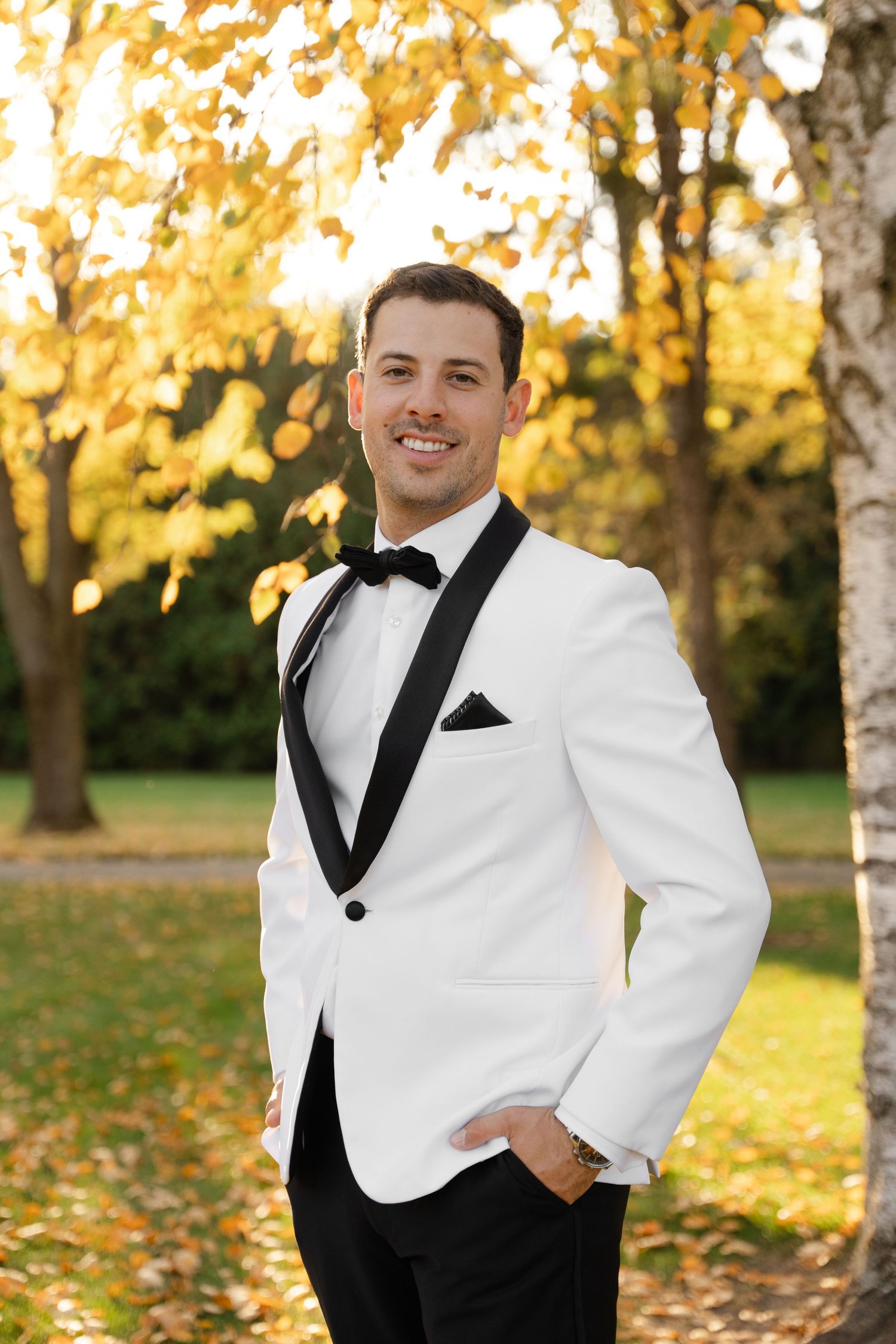 Groom smiles in classic white dinner jacket and black bow tie during outdoor autumn wedding.