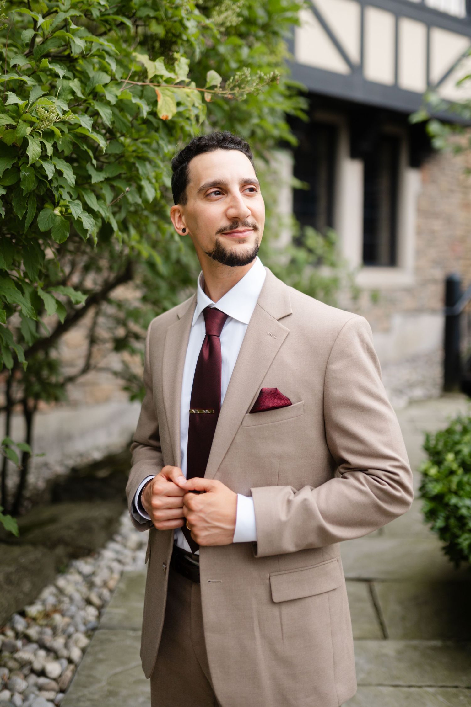 Professional in light colored suit poses outdoors against stone building and greenery.