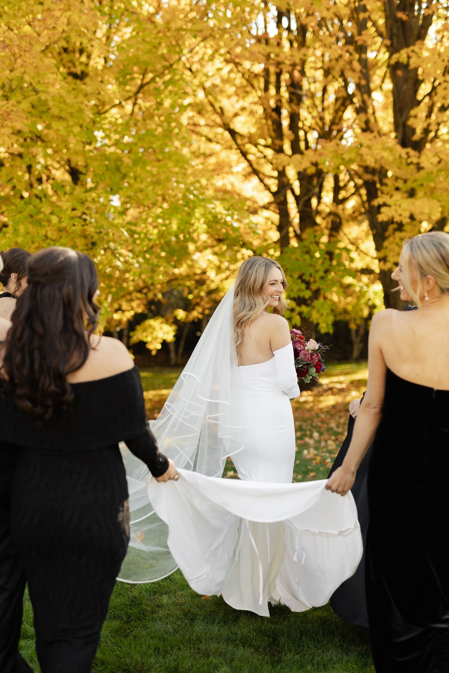 Bridesmaids in black helping with bride's dress against golden fall trees.